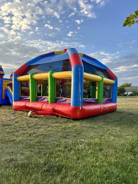 Colorful inflatable bounce house with red, blue, yellow, and green sections on a grassy field under a partly cloudy sky.