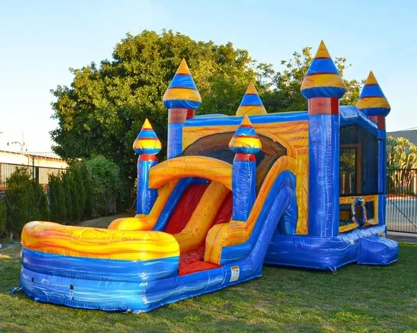 Colorful inflatable castle with a slide, set outdoors on grass with trees and a fence in the background.