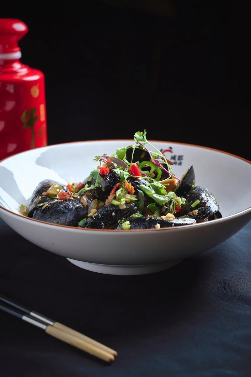 A bowl of black mussels garnished with chopped green onions, red chili peppers, and microgreens, with a pair of chopsticks and a red container in the background.