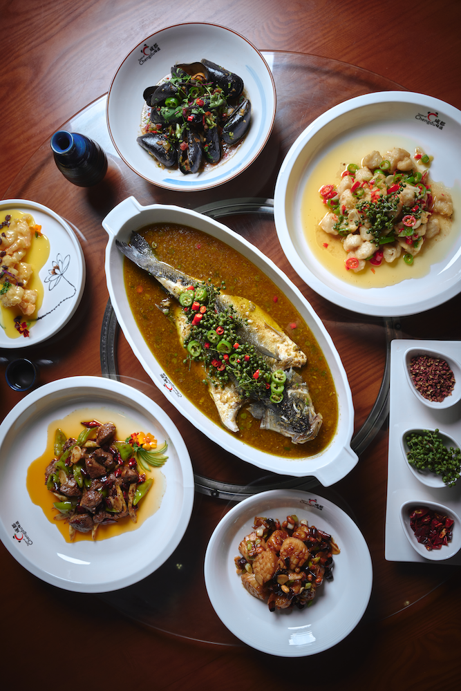 A traditional Chinese meal featuring a variety of dishes on a wooden table, including steamed fish in sauce, shrimp in sauce, stir-fried vegetables with meat, stir-fried chicken, and black mussels garnished with herbs and peppers, with small bowls of spices on the side.