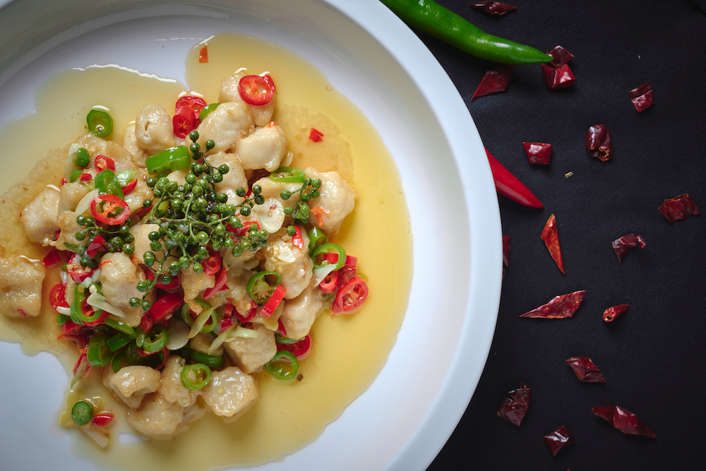 A plate with fish topped with chopped red and green chili peppers and green peppercorns. Green and red chili peppers and crushed dried red chili peppers are scattered around the plate on a black surface.
