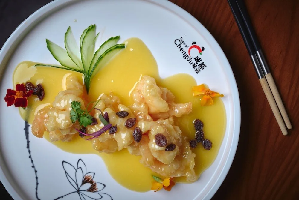 Shrimp with orange liquor sauce, garnished with cucumber slices, edible flowers, and a sprig of cilantro, served on a white plate with a black floral design.