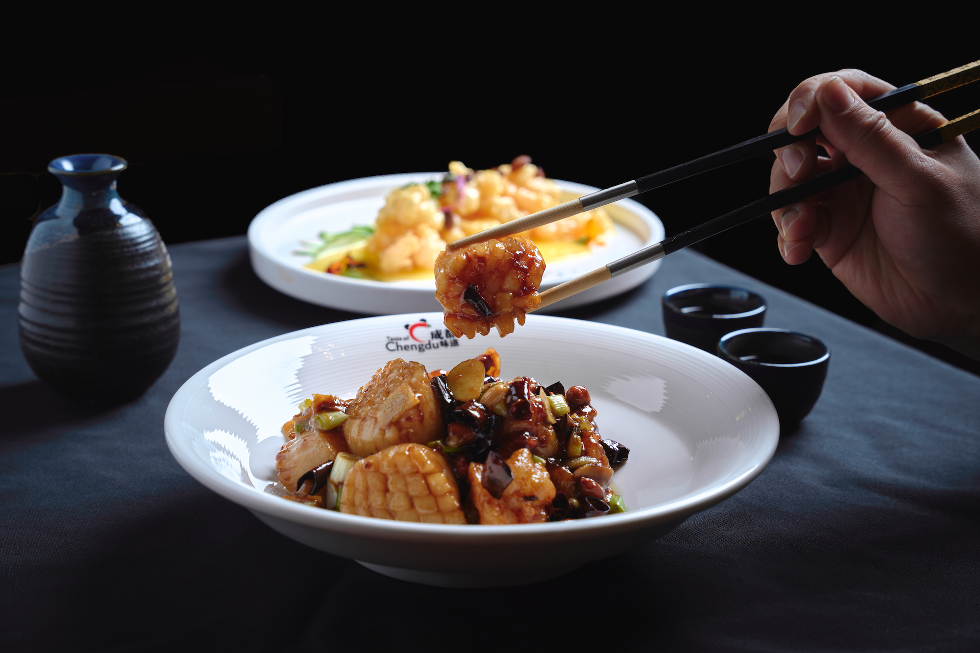 Close-up of a hand holding chopsticks picking up a scallop from a white plate . In the background, there is another white plate with shrimp a small blue vase, and small black bowls on a black surface with a black background.
