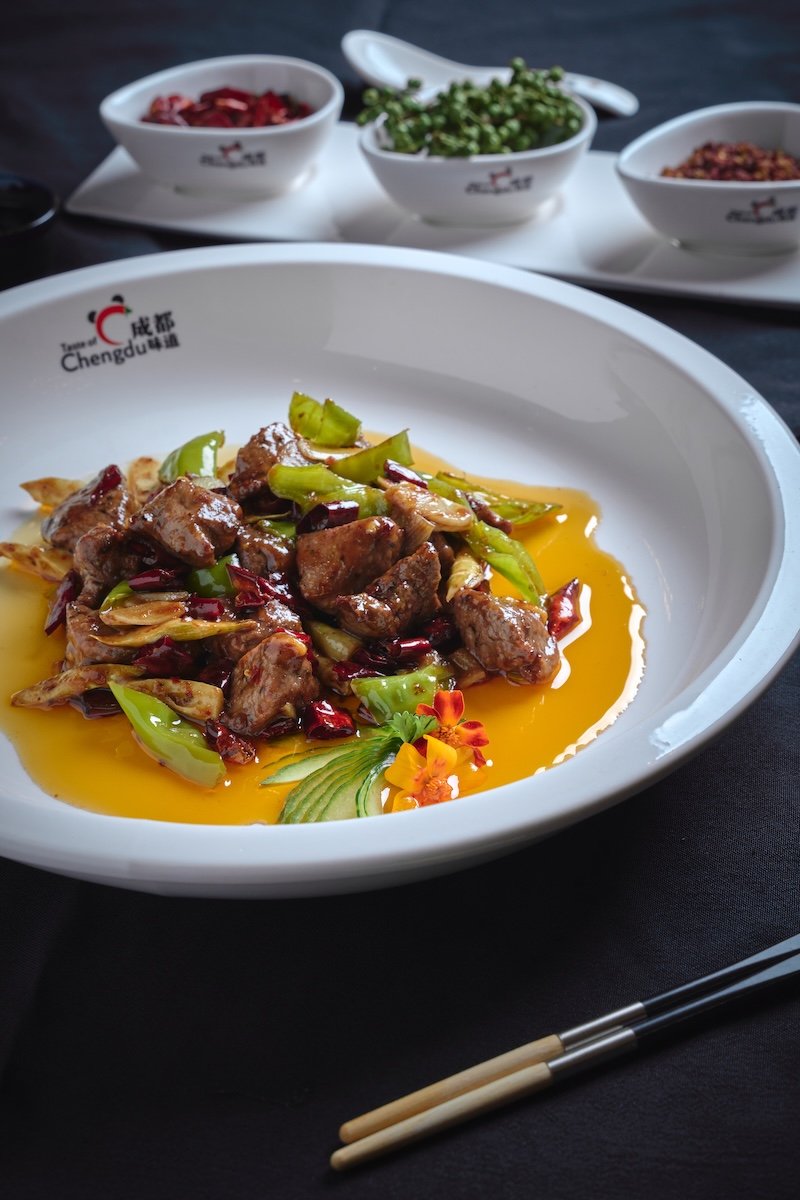 A plated dish of filet mignon with green vegetables and red chili slices, garnished with a flower and herbs, served on a white Chengdu-branded plate, with three small bowls of sauces and herbs in the background.