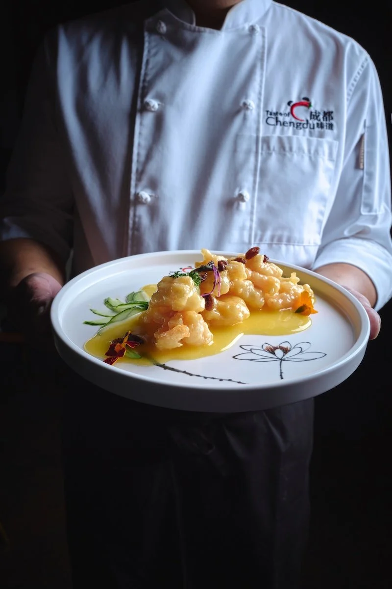 Chef Tiger in white uniform holding a white plate with shrimp topped with orange liquor sauce and garnished with herbs and vegetables.