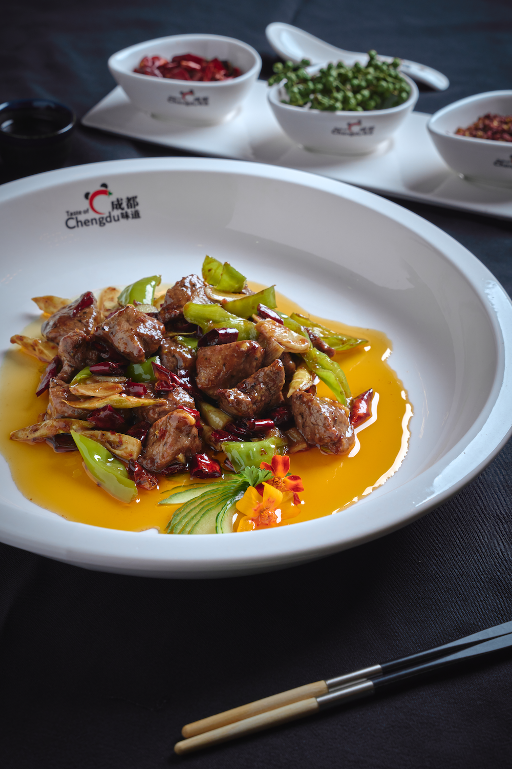 A plate of Filet Mignon with vegetables and sauce, garnished with flowers and cucumber slices, with small bowls of condiments and chopsticks on a black table.