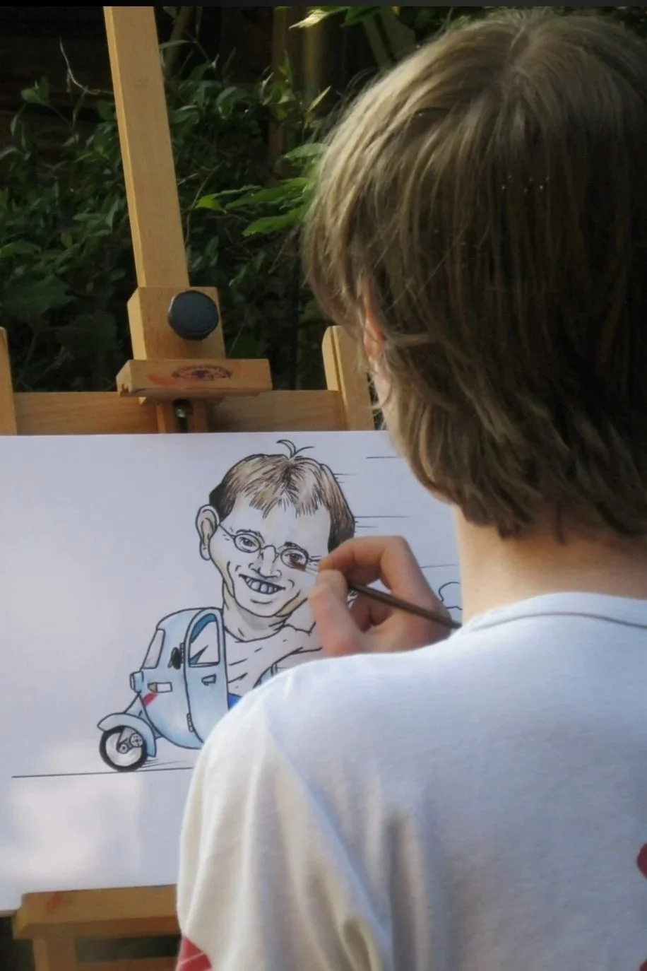A person drawing a caricature of a smiling man with glasses on a white paper, with a small three-wheeled vehicle illustrated next to the caricature.