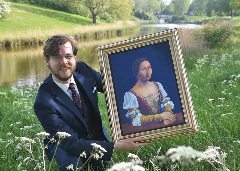 A man in a suit holding a framed portrait of a woman in historical clothing outdoors near a river and green field.
