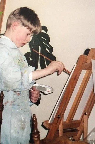 A young boy painting on an easel indoors, holding a paintbrush and palette.