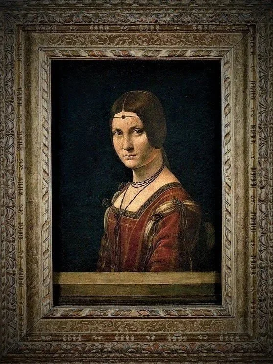 A portrait of a young woman with dark hair, wearing a brown dress with puffed sleeves and multiple necklaces, framed in an ornate wooden frame.