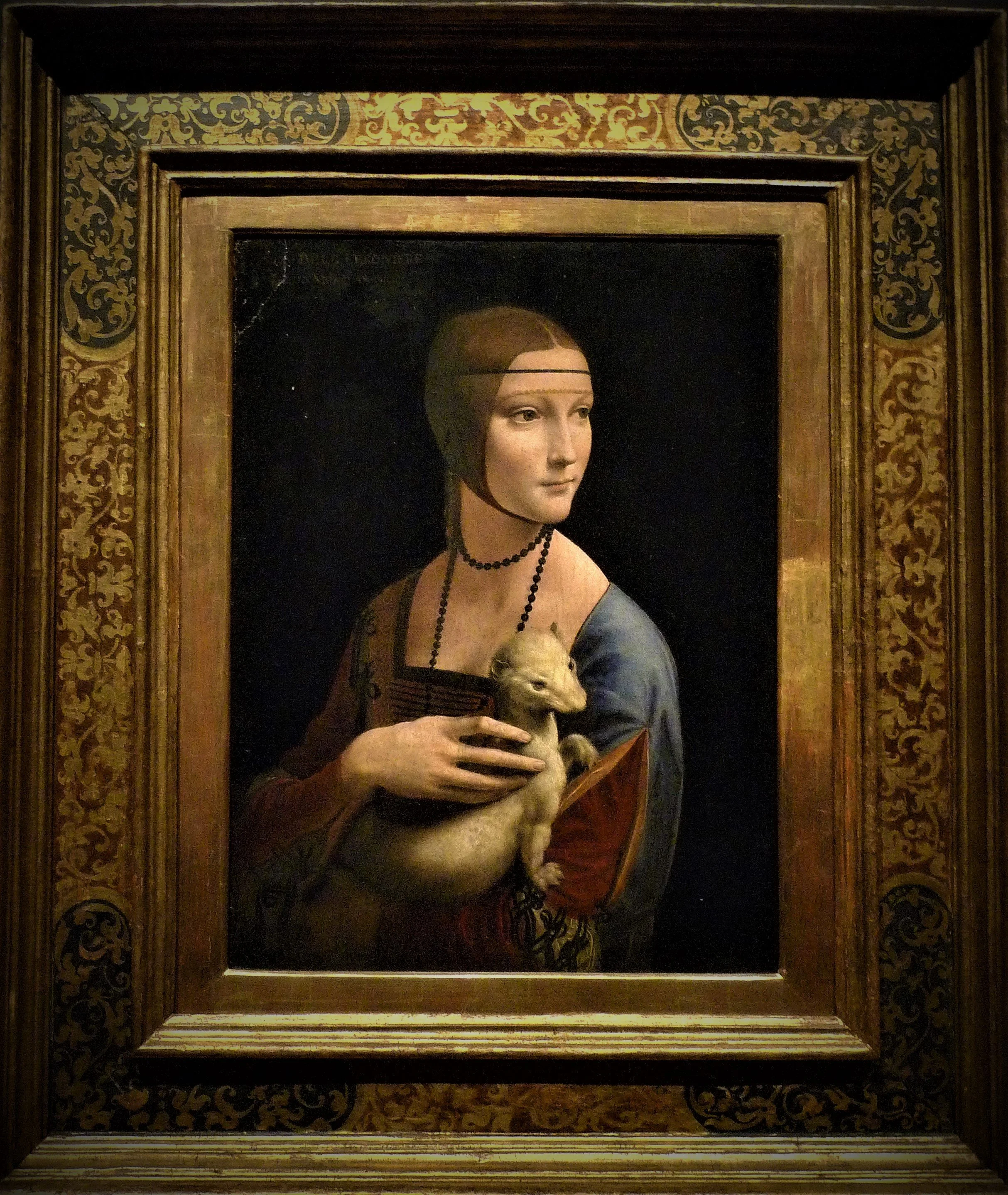 A painting of a woman holding a small dog, framed with ornate gold and dark brown wood, displayed on a wall.