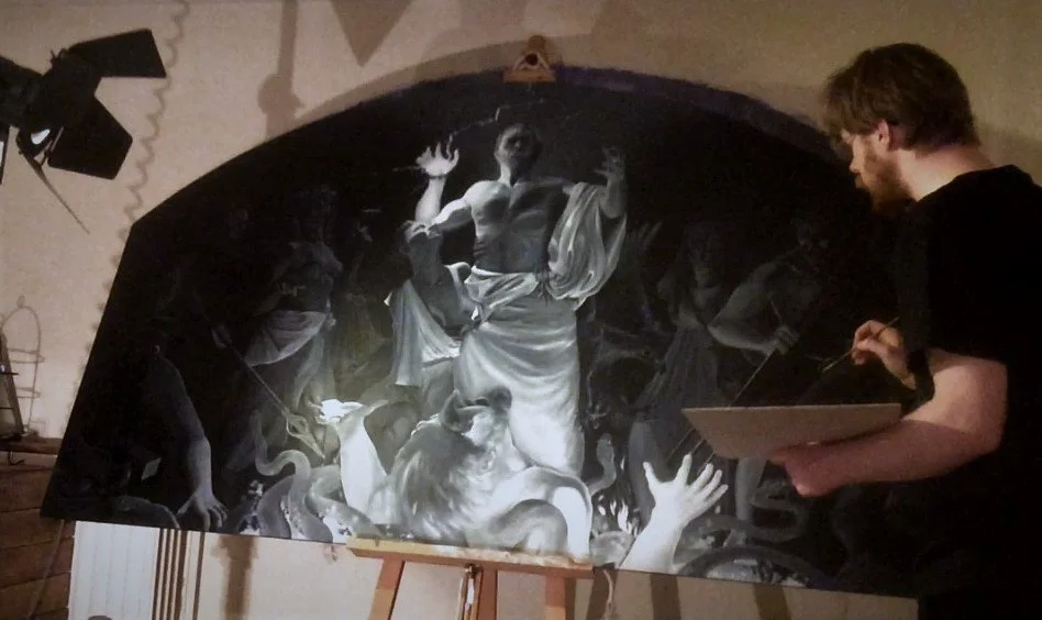 A man with glasses and dark hair holding a tablet, observing a large grayscale painting on an easel, depicting multiple figures, including a central woman with her arms raised and a lion, in a dramatic scene.
