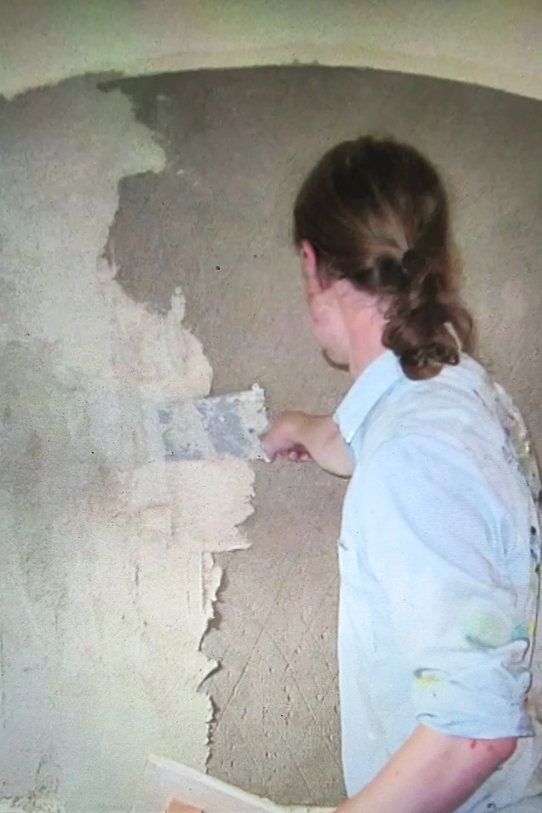 Person with brown hair in a bun repairing a damaged wall by applying plaster or joint compound.