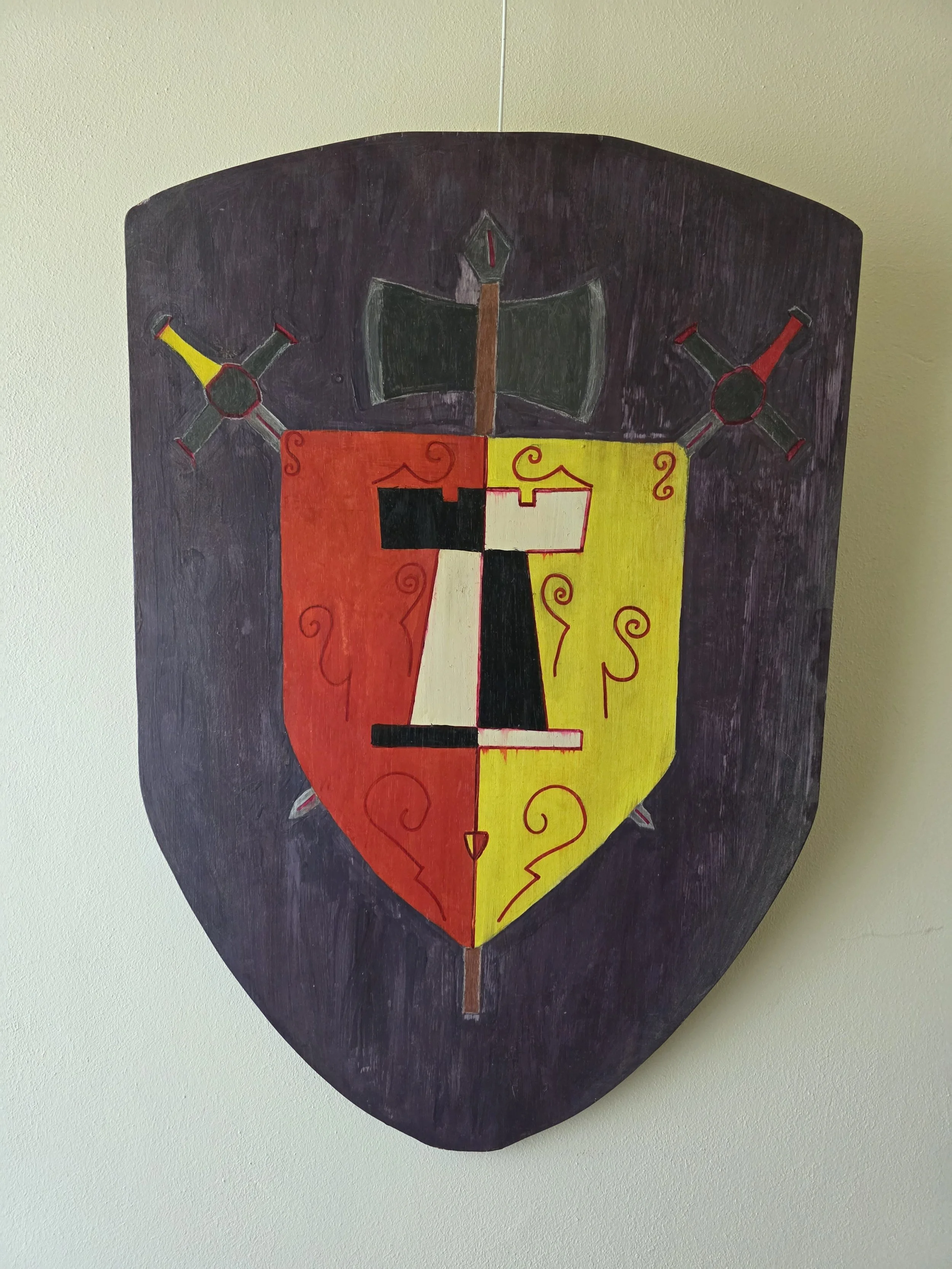 A wooden shield with a painted heraldic design divided into two colors, red and yellow, with a black and white section in the center. The shield features a black axe at the top and two crossed hammers or tools with red and yellow ends.