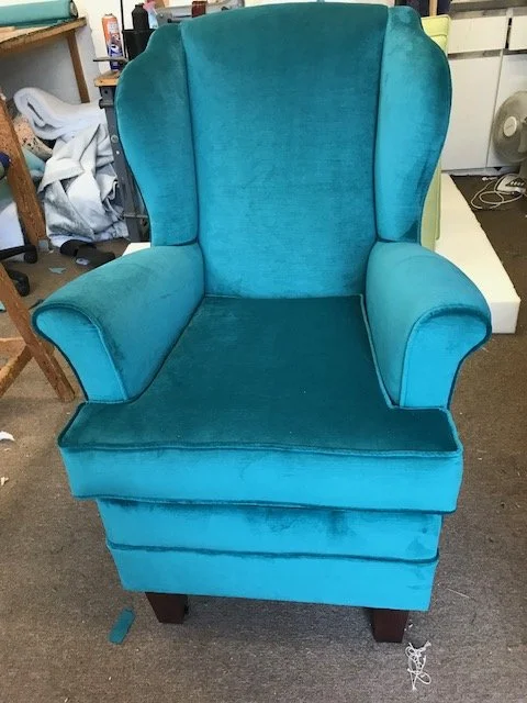 A vintage turquoise armchair with a high back and rolled arms, sitting on a brown carpeted floor in a cluttered room.