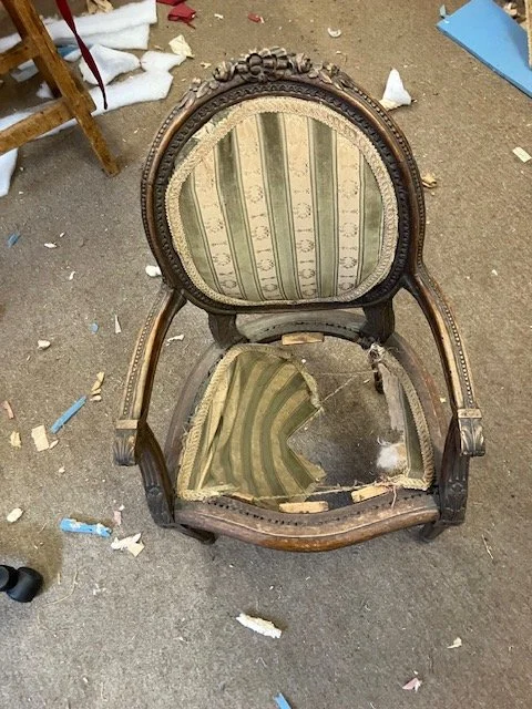 A broken antique wooden chair with upholstered backrest and seat, both torn and damaged, surrounded by debris on the floor.