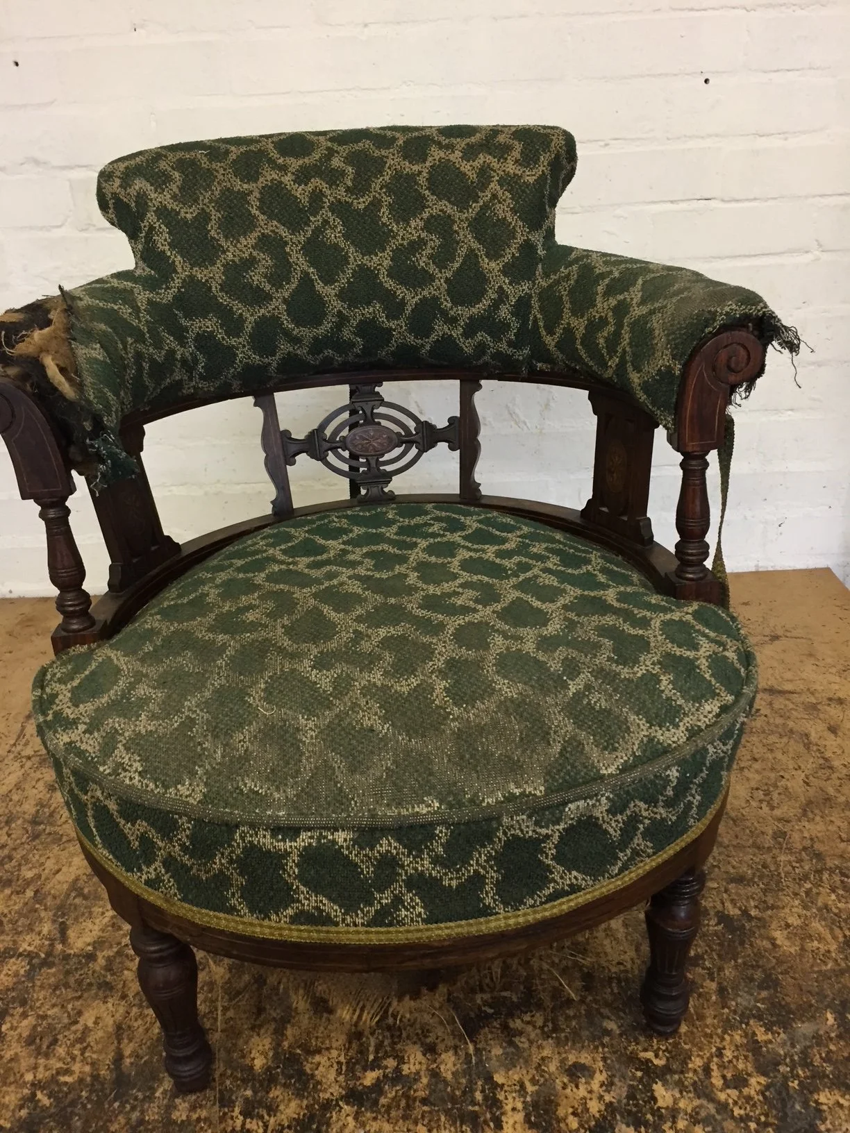 An antique wooden armchair with green patterned upholstery, featuring a damaged armrest and decorative metalwork in the backrest, set against a white brick wall.