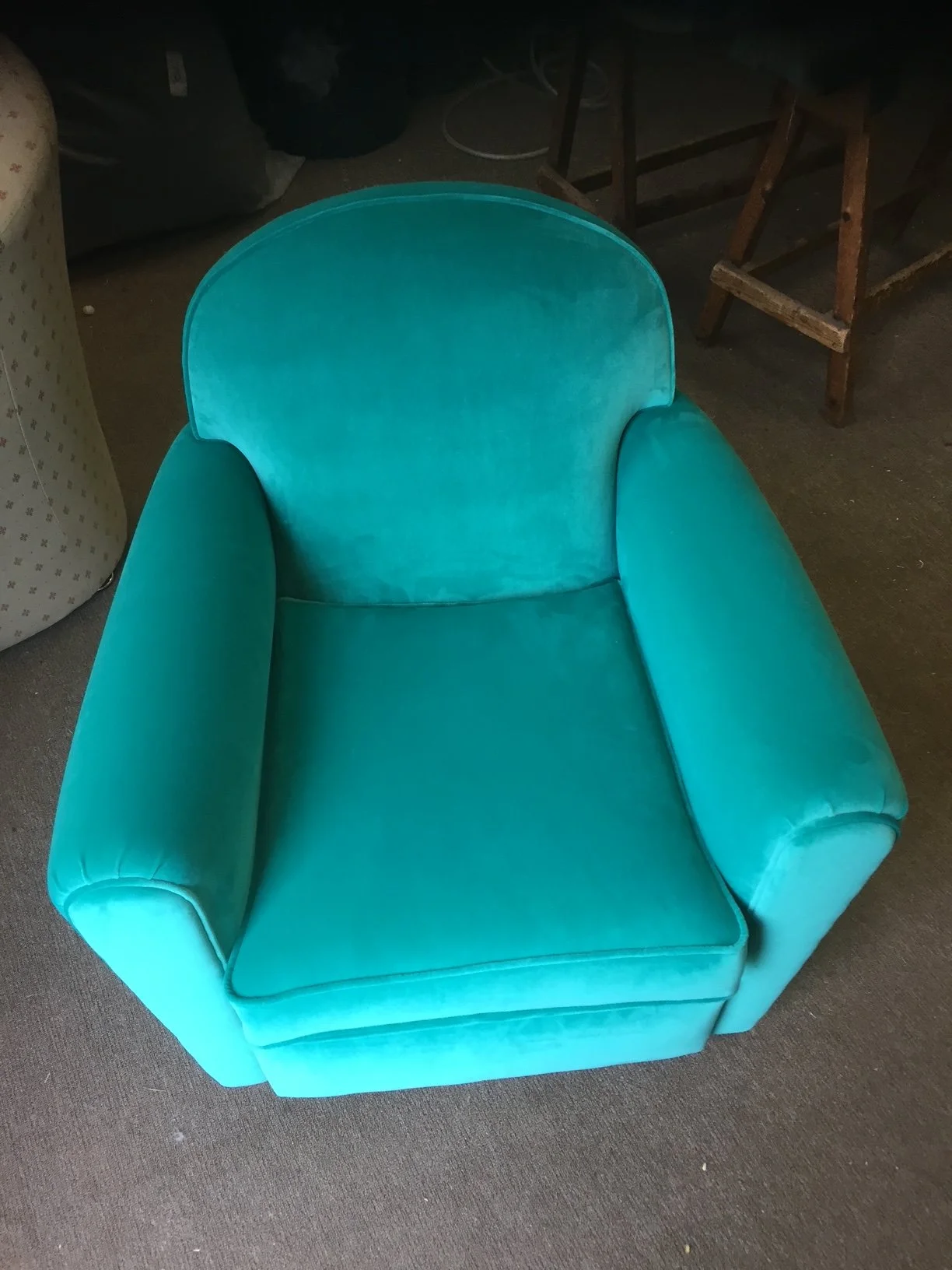 A vintage turquoise armchair with curved armrests and a rounded back, sitting on a brown carpeted floor. In the background, there is a wooden step stool and some miscellaneous items.