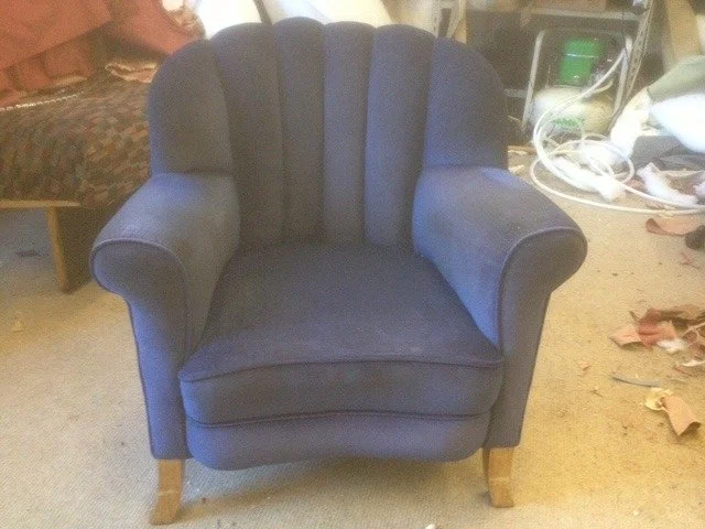A blue upholstered armchair with a rounded back and wooden legs in a cluttered room.