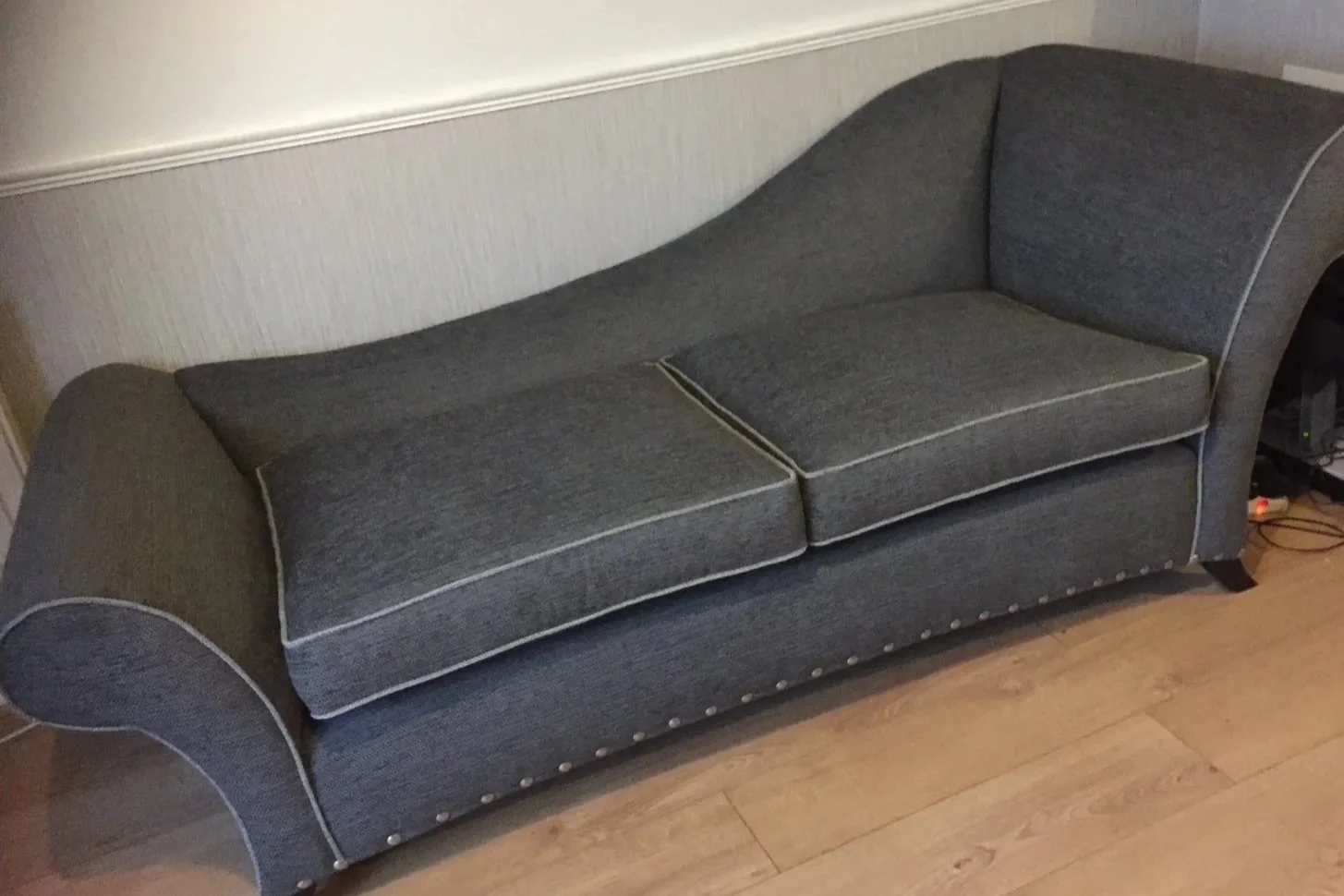 A grey fabric sofa with rolled arms and decorative trim along the edges, positioned against a wall on a wooden floor.
