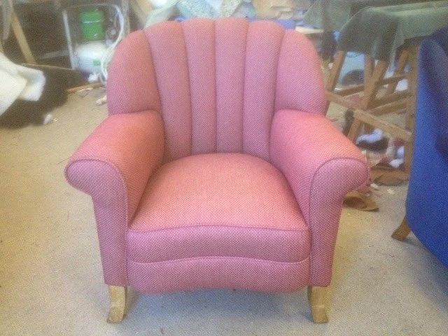 Pink upholstered armchair with wooden legs in a cluttered room.