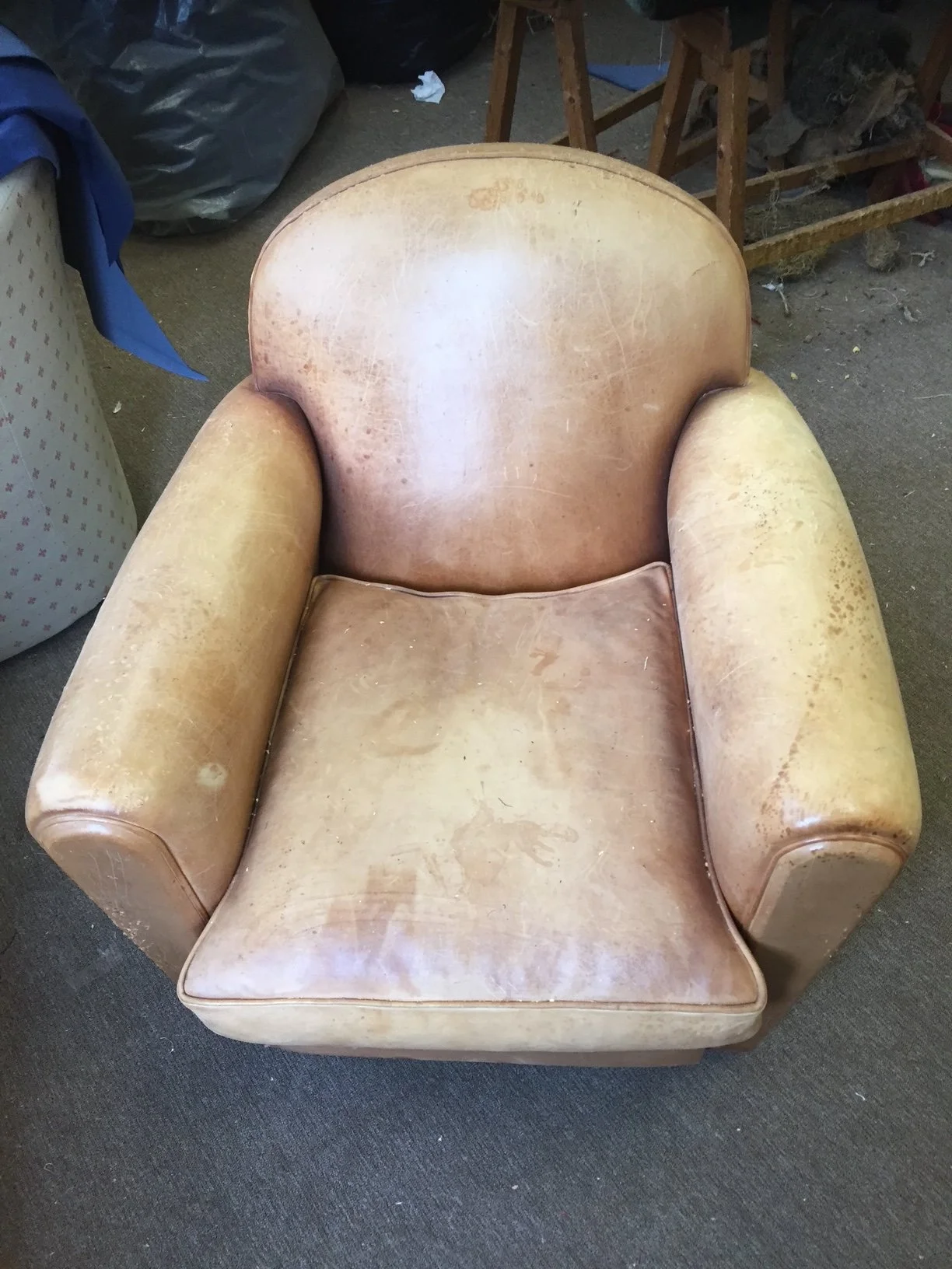 An old, worn yellowish armchair with a cracked and stained leather surface, sitting on a dark carpeted floor, surrounded by miscellaneous items and wooden furniture.