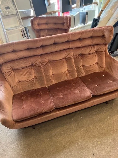 Brown velvet upholstered sofa with tufted backrest and cushions, set in a room with windows, shelves, and other furniture.