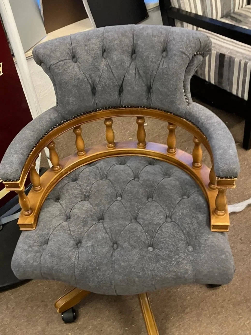 A grey upholstered office chair with tufted backrest and seat, featuring wooden armrests and spindles, and black caster wheels.