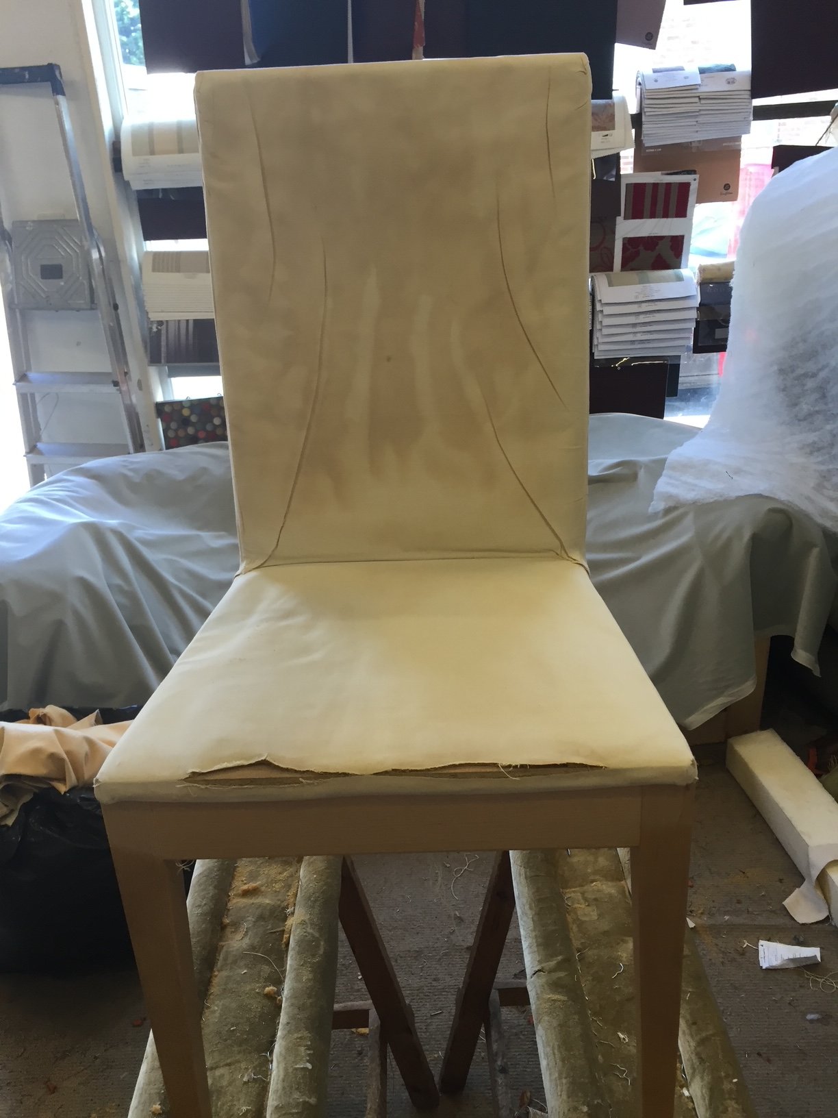 A partially upholstered yellow chair with visible seams and worn edges, placed on a wooden sawhorse in a workshop surrounded by boxes, fabrics, and tools.