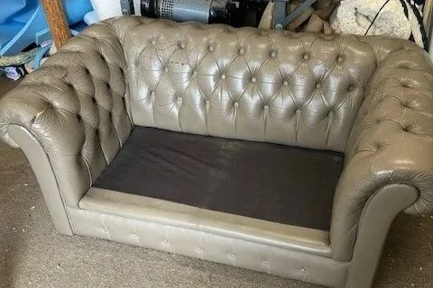 A beige tufted leather couch with rolled arms in a cluttered storage area.