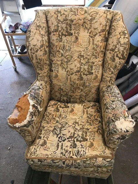 An old, worn upholstered armchair with floral fabric, torn on the arms revealing foam and stuffing, in a cluttered room.