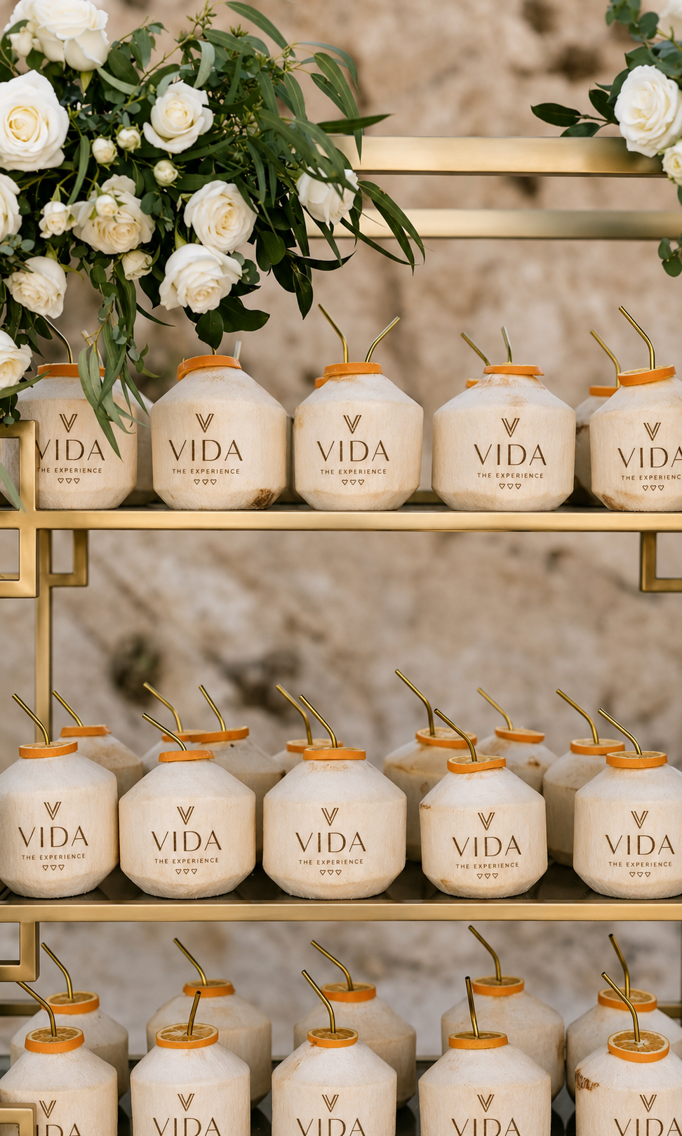 A display of coconut drinks with straws, arranged on golden shelves, with a bouquet of white roses and greenery on the left side.