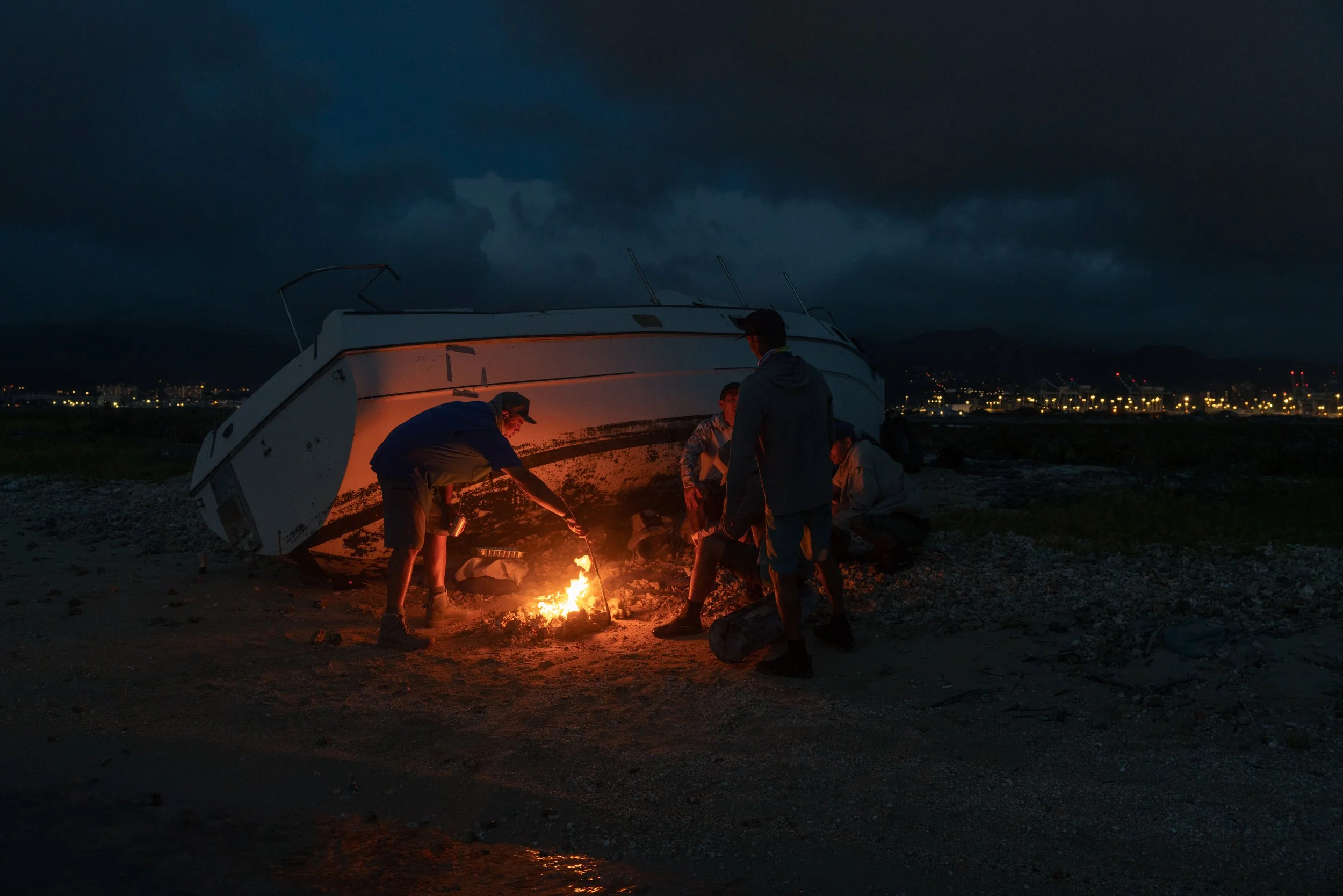 People gathered around a small campfire outdoors at night, with a partially sunken boat and city lights in the background.