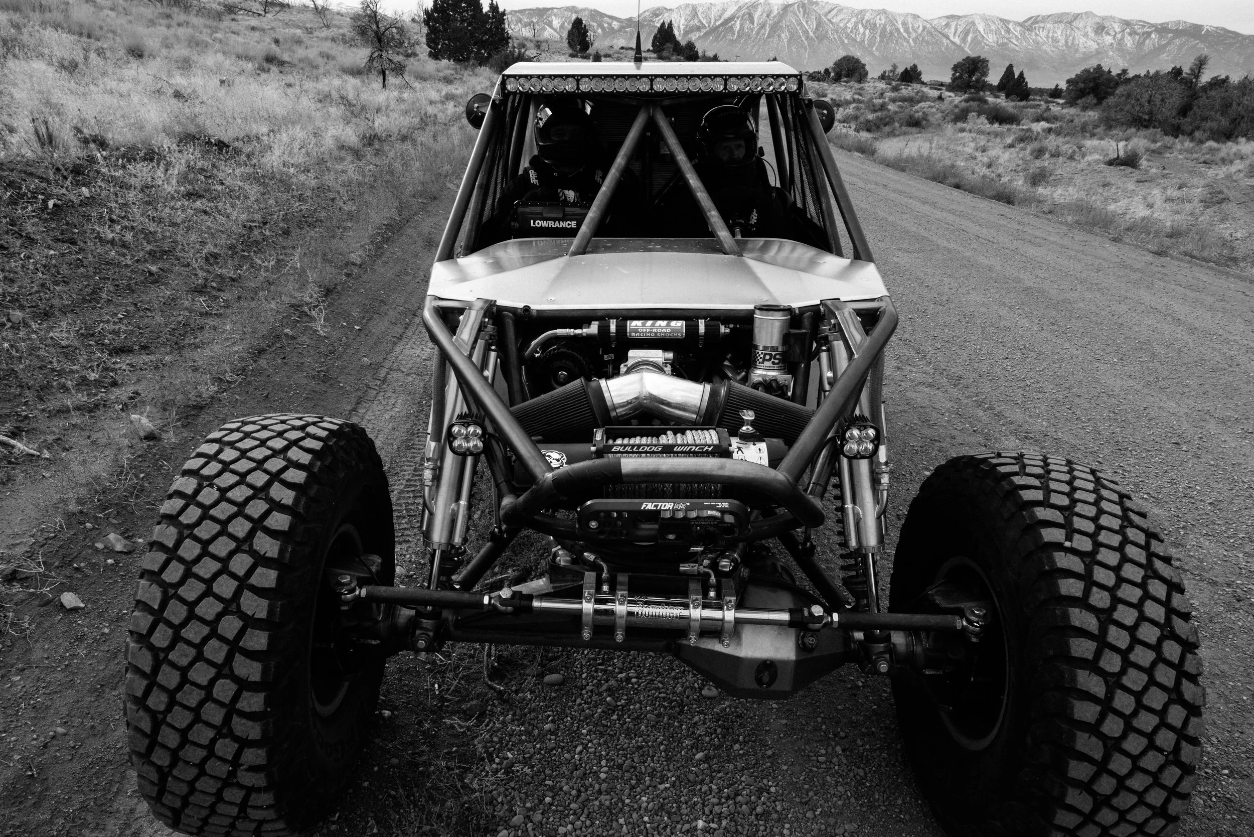Black and white photo of an off-road dirt vehicle with large tires on a dirt road in a rural area, mountains in the background, and two people wearing helmets inside.