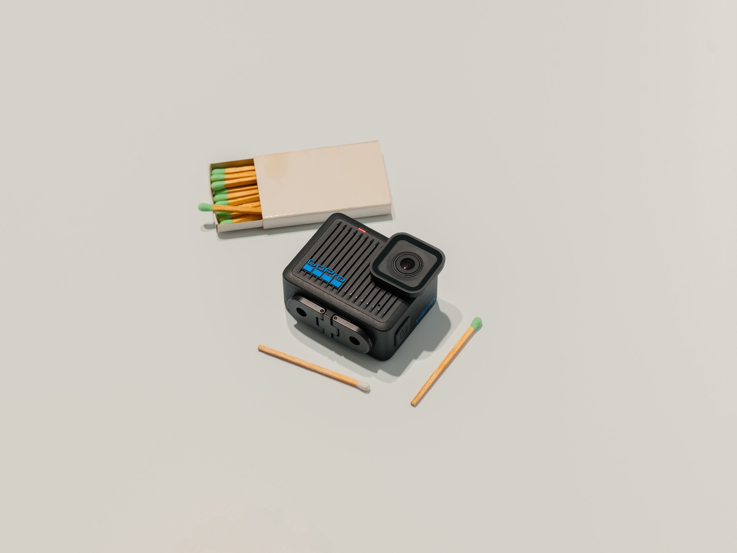 Close-up of a white matchbox containing green-tipped matches, partially opened, on a plain, light-colored surface. A black GoPro camera is positioned nearby, with one wooden matchstick lying flat in front of it.