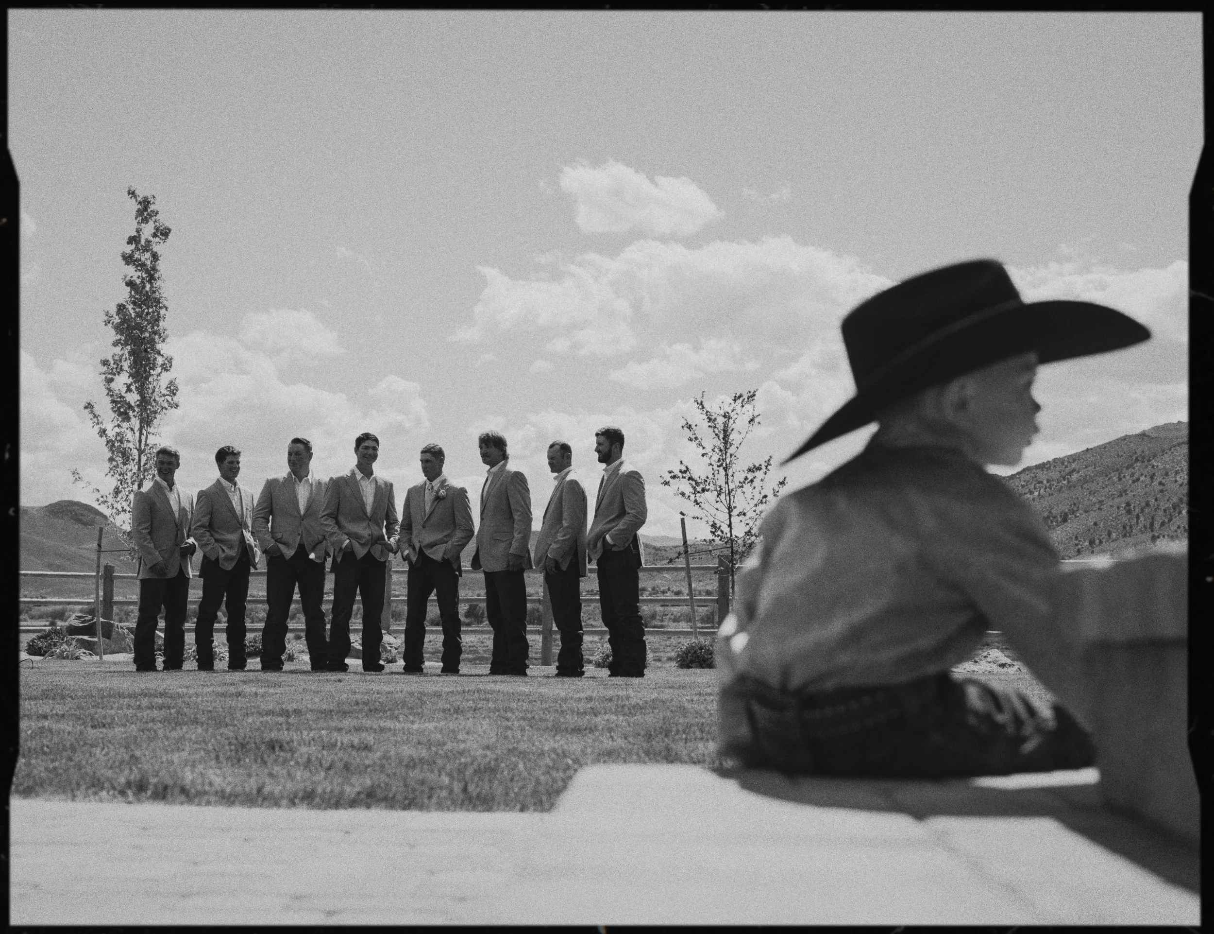 A man in a cowboy hat sitting in the foreground with a group of men in suits standing in a line behind him outdoors under a partly cloudy sky.