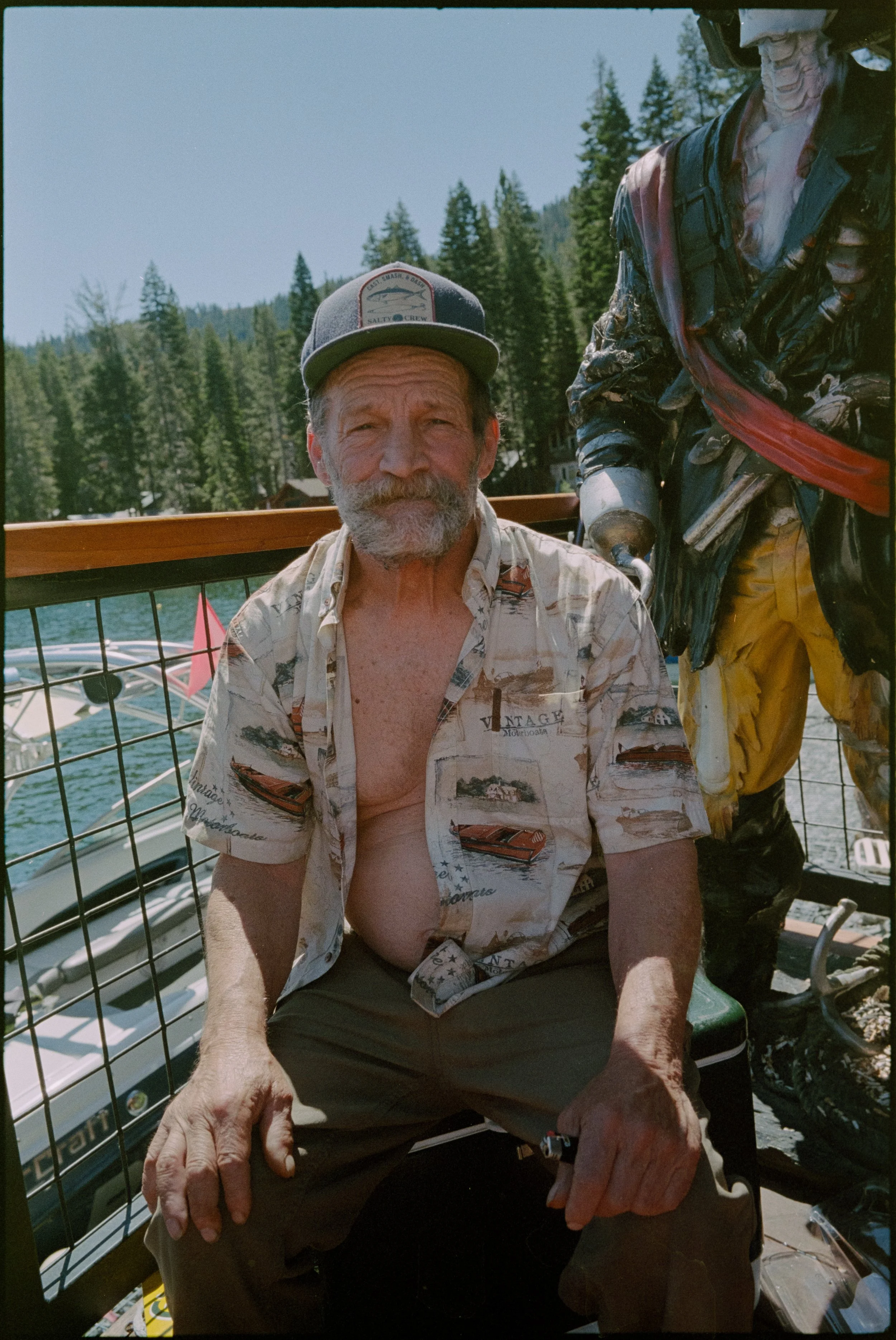 An elderly man with a gray beard and mustache, wearing a gray cap with a patch, sitting outdoors on a boat, with a boy's sailboat, trees, and mountains in the background.