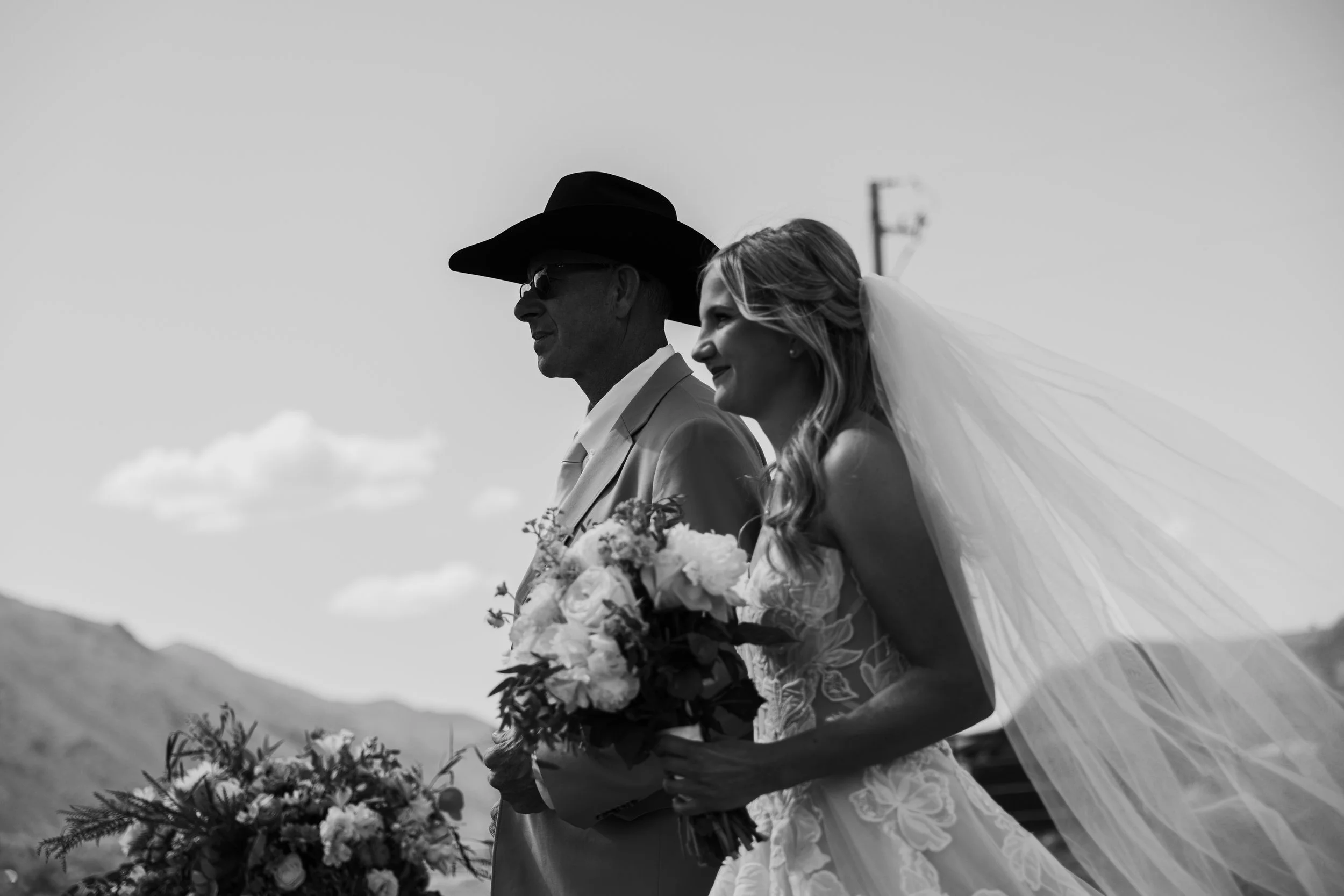 A black and white photo of a bride and an older man, possibly her father, standing outdoors, both smiling. The bride is wearing a wedding dress and veil, and holding a large bouquet of flowers. The man is wearing sunglasses, a suit, and a cowboy hat.