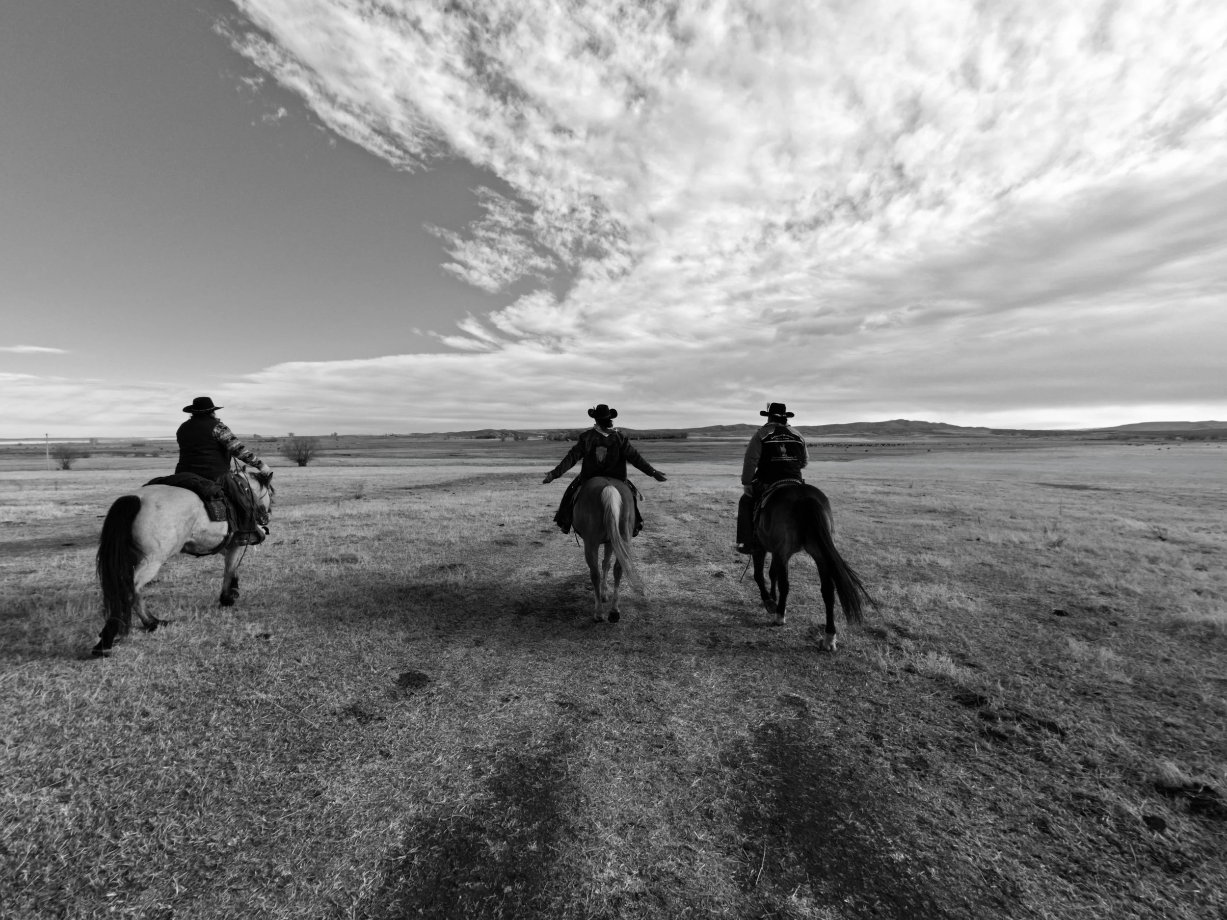 Three cowboys riding horses across an open plains landscape under a partly cloudy sky