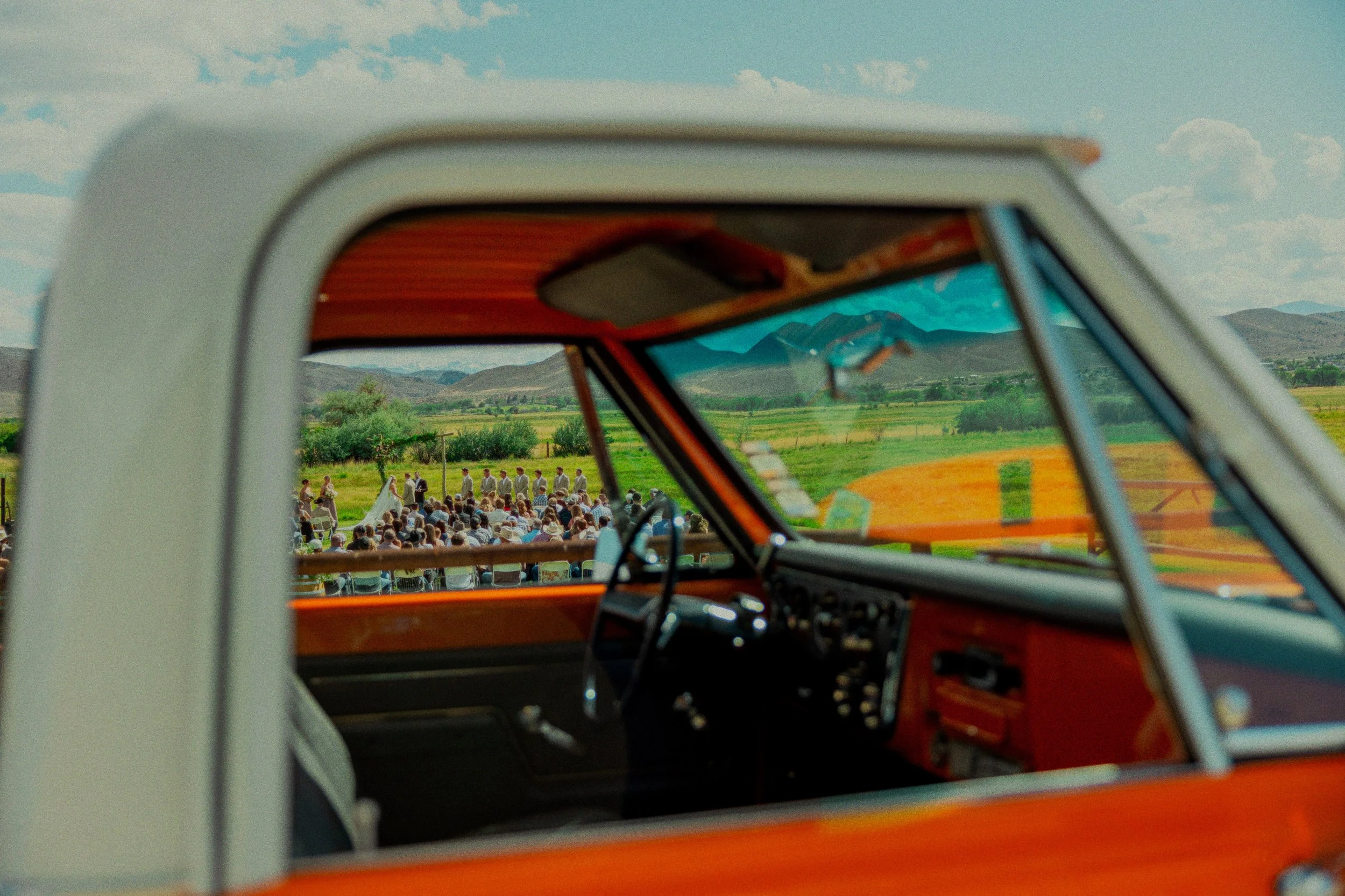 The image shows the interior of an orange vintage van with a view through the windshield of a large outdoor gathering of people sitting on chairs in a green field, with mountains and a partly cloudy sky in the background.