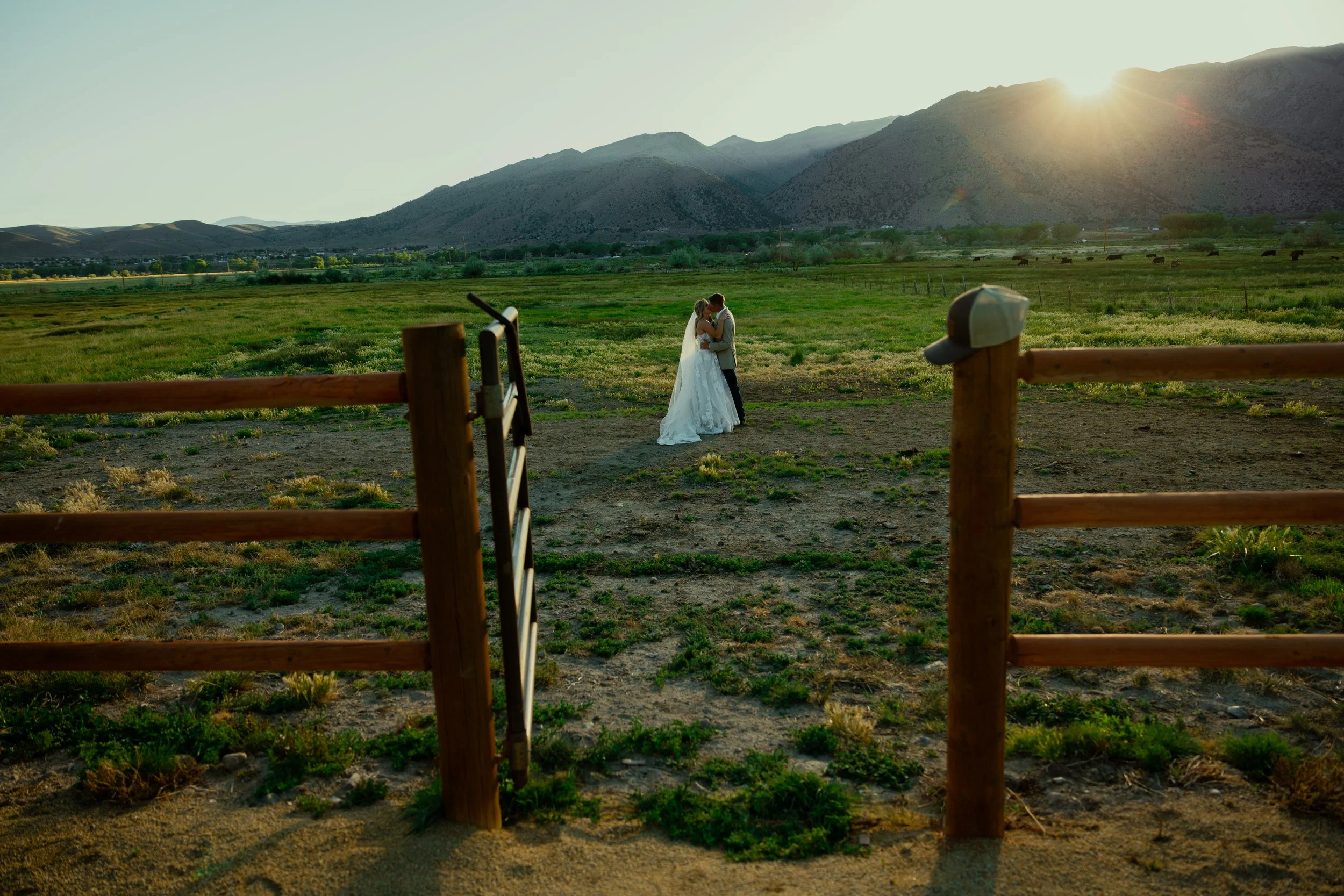 A bride and groom embracing and kissing in a grassy field at sunset, with mountains in the background and a wooden fence in the foreground.