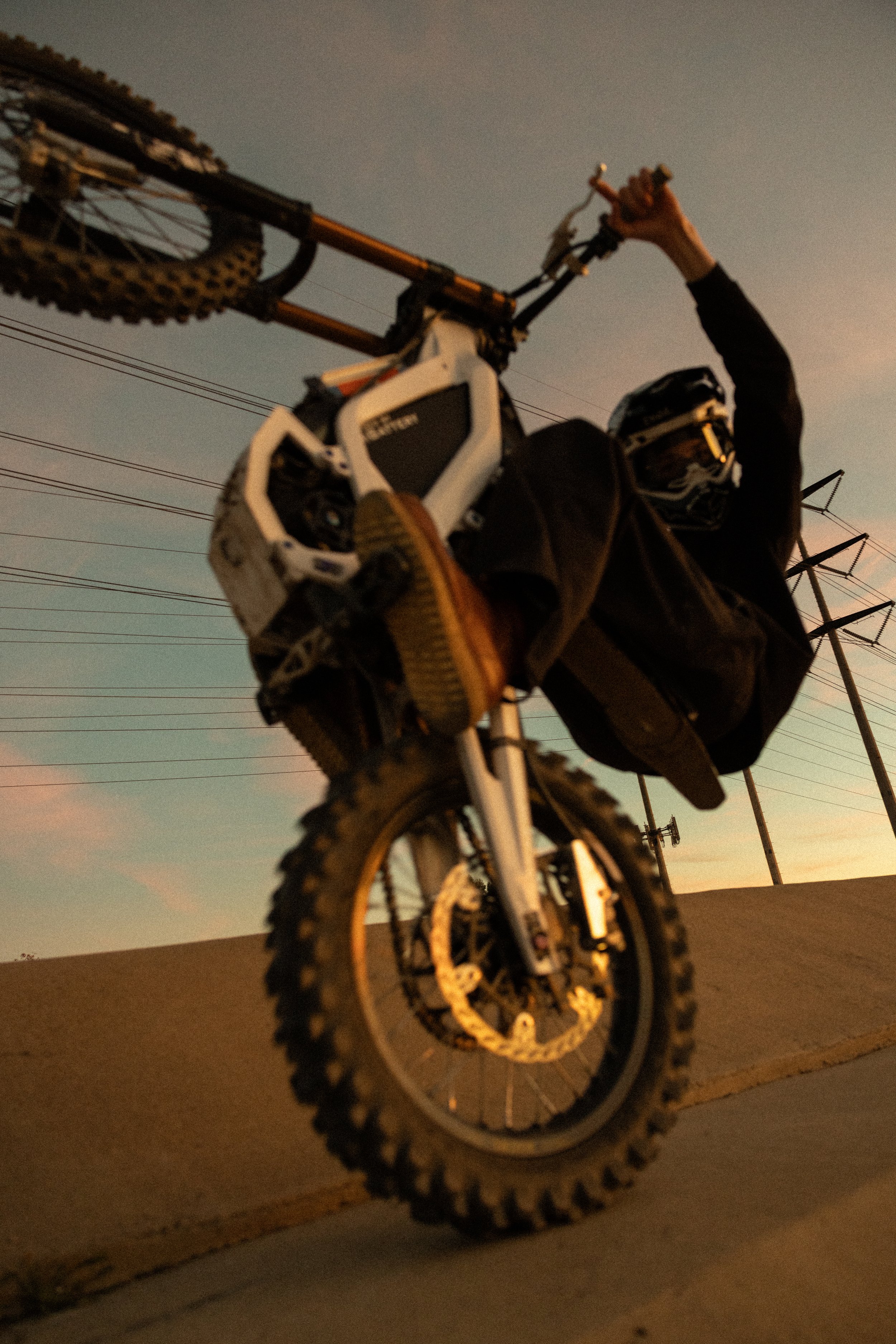 A person wearing a helmet and black clothing performing a wheelie on a dirt bike during sunset with power lines in the background.
