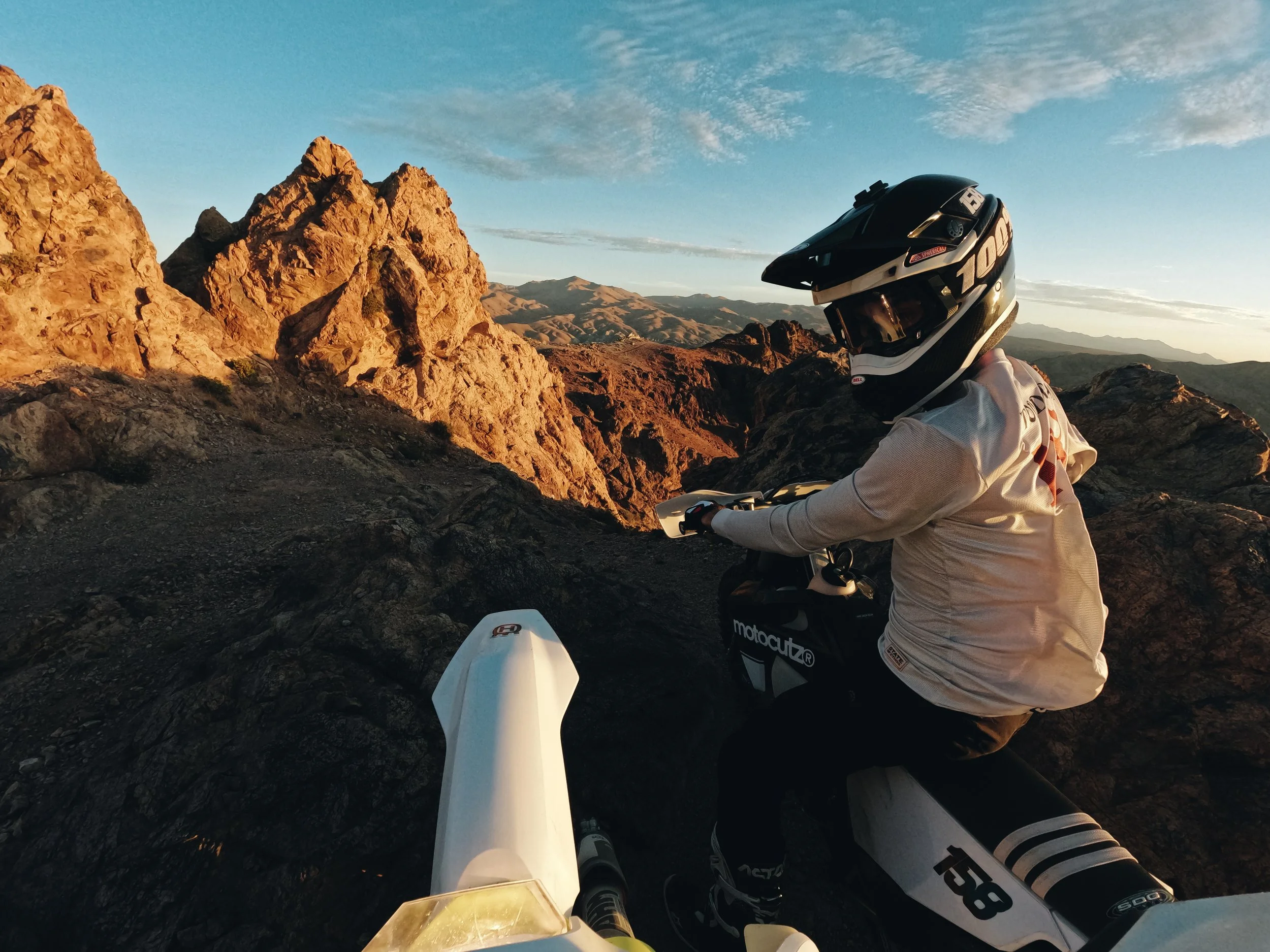 A motorcyclist wearing a helmet and gloves riding through a rocky desert landscape during sunset or sunrise.