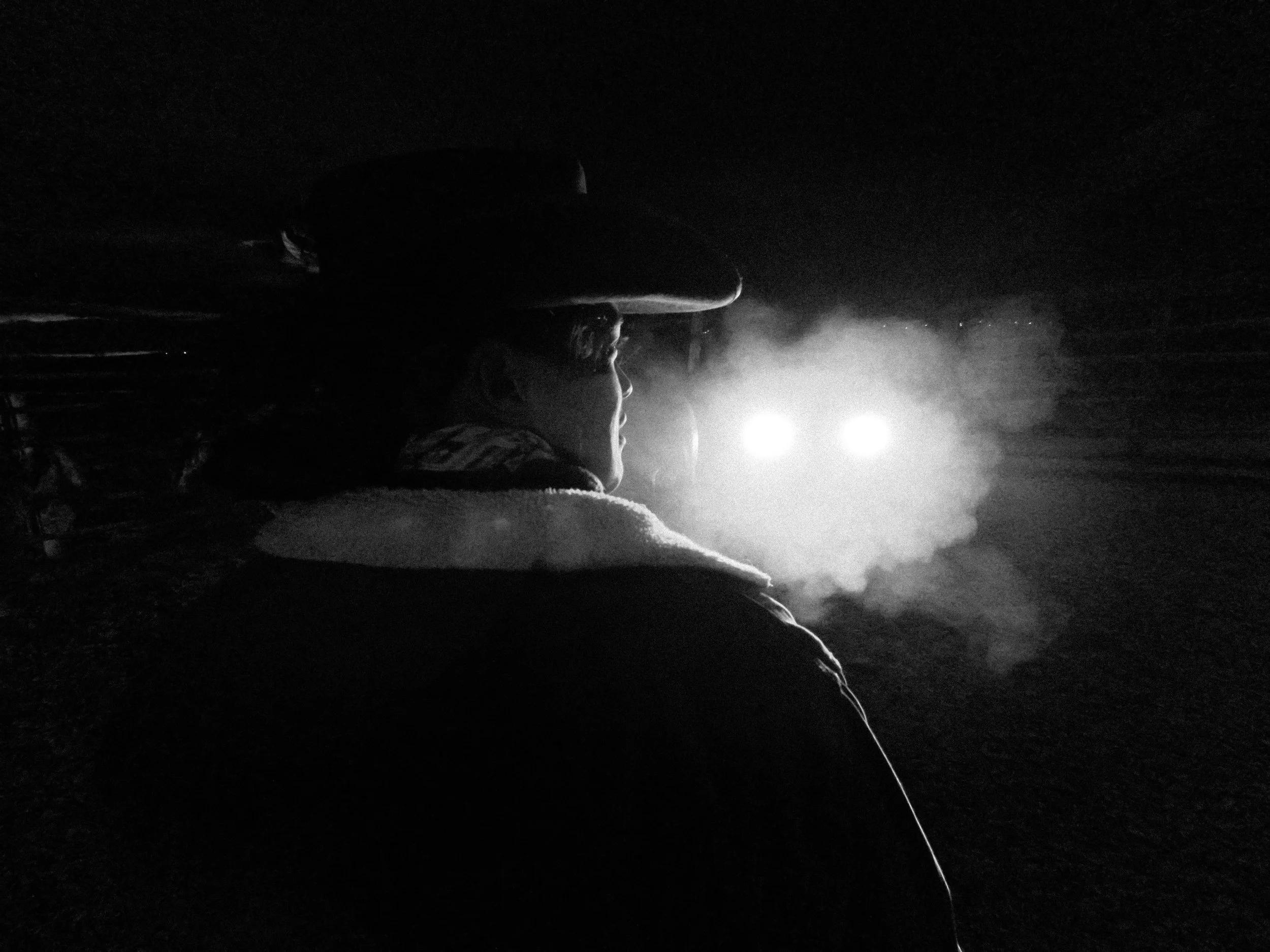A person wearing a hat is seen in profile, illuminated by a bright light source in a dark environment, with steam or smoke in the background.