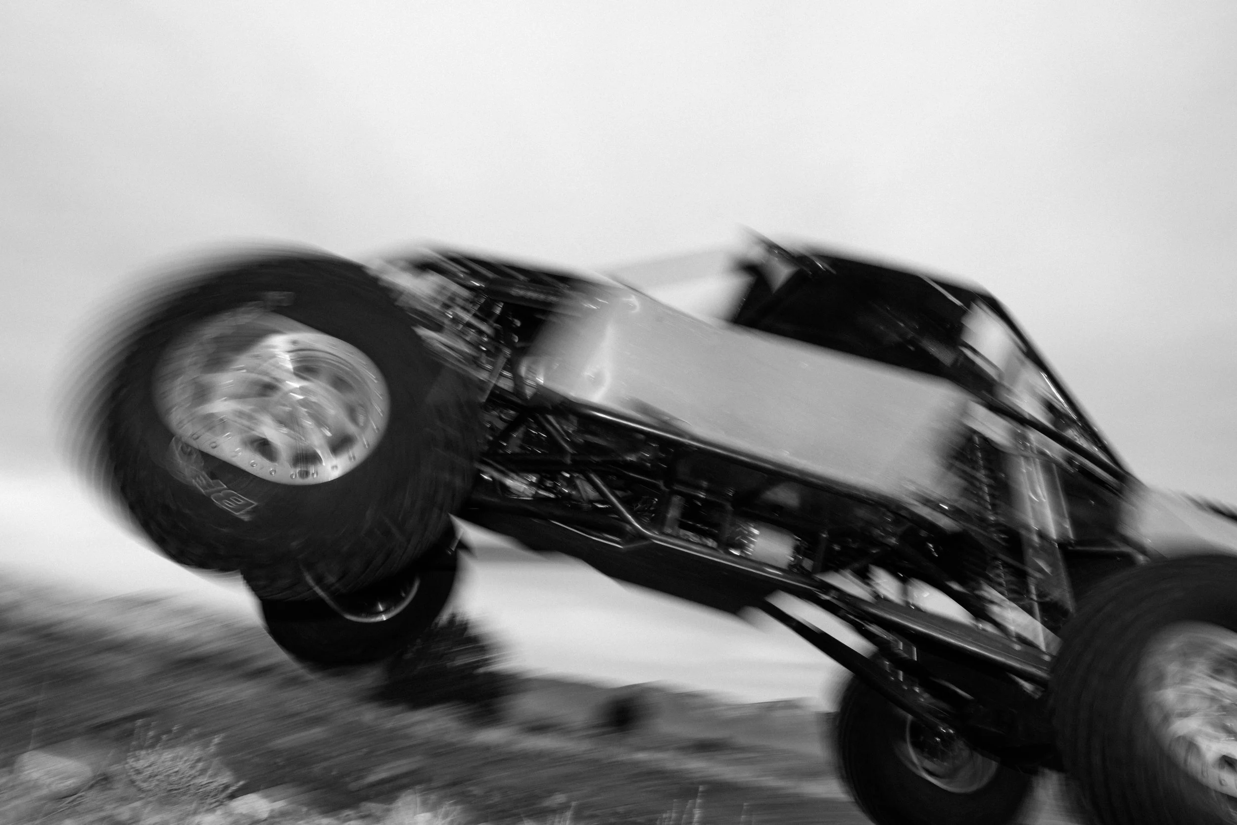 A black and white photograph of a race car in motion, with blurred wheels and surrounding environment.