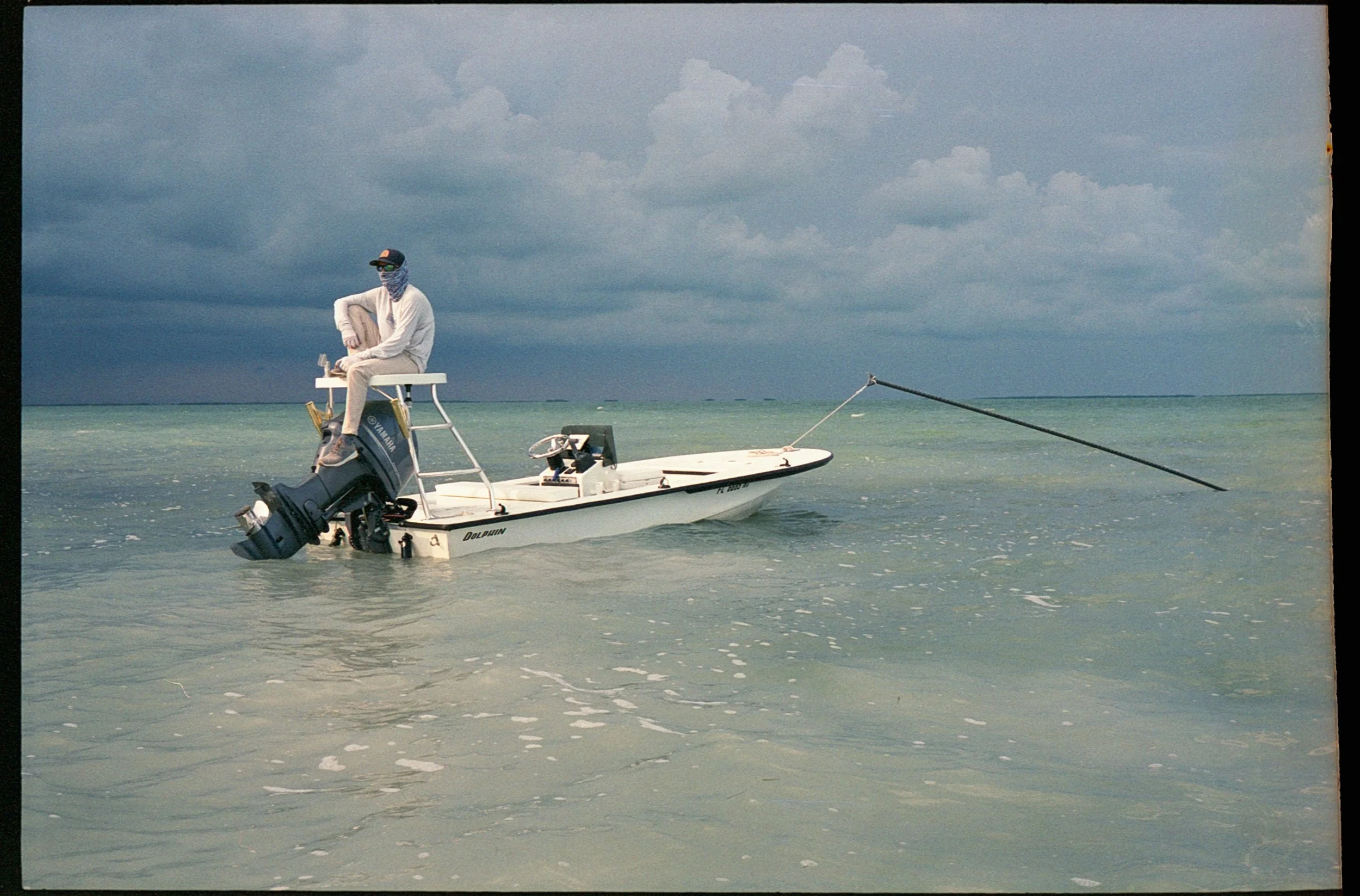 A person sitting on the edge of a small white boat with a Yamaha outboard motor, in calm ocean waters under a cloudy sky, holding a fishing rod.