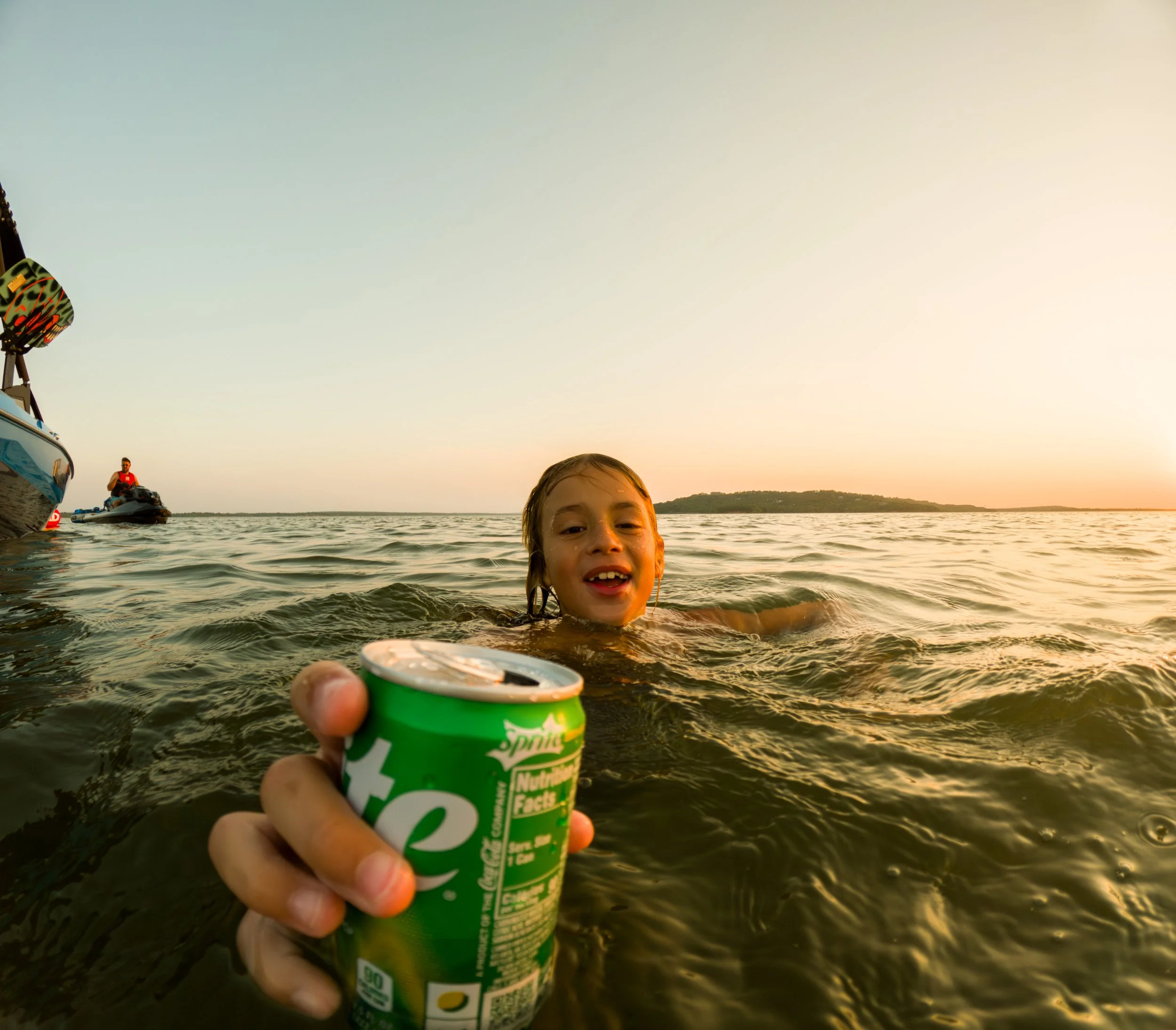 A child swimming in a body of water during sunset, holding a green can of Sprite in the foreground, with boats and a person sitting on a boat in the background.