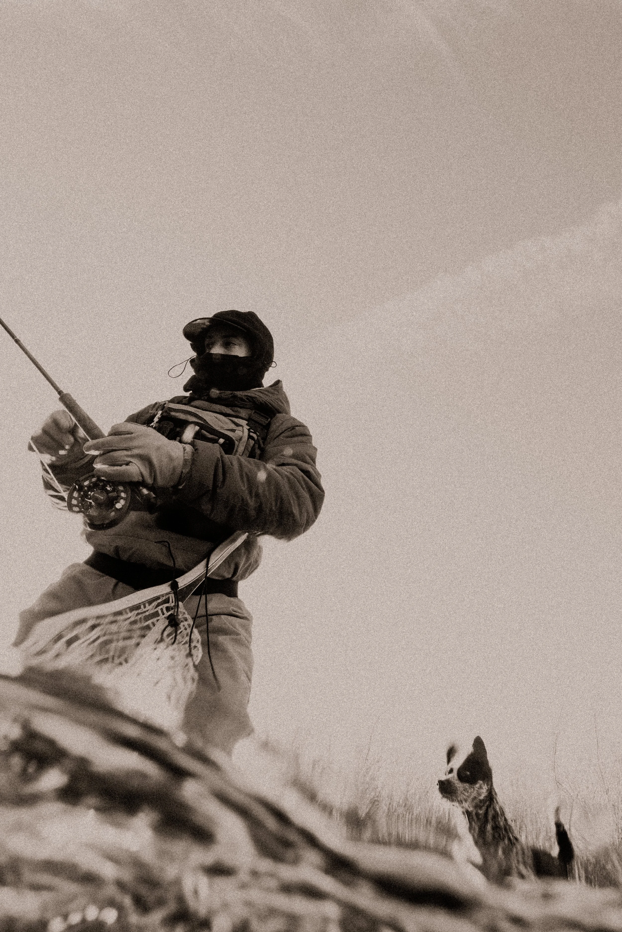 A person wearing winter clothing, including a hat and a face covering, is holding a fishing rod and sitting on the snow. A dog is visible in the background, looking at the camera, in a snowy outdoor setting.