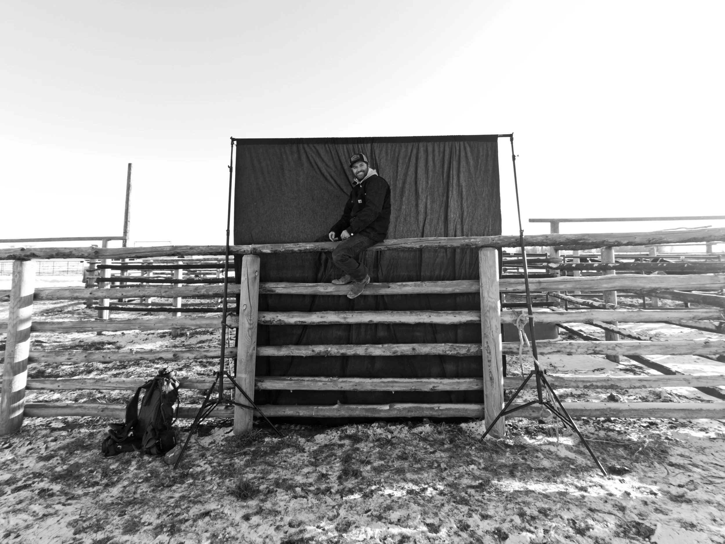 A man with a beard and a cap sitting on a wooden railing in front of a black cloth backdrop, with equipment like tripods and a backpack in an outdoor setting with snowy ground.