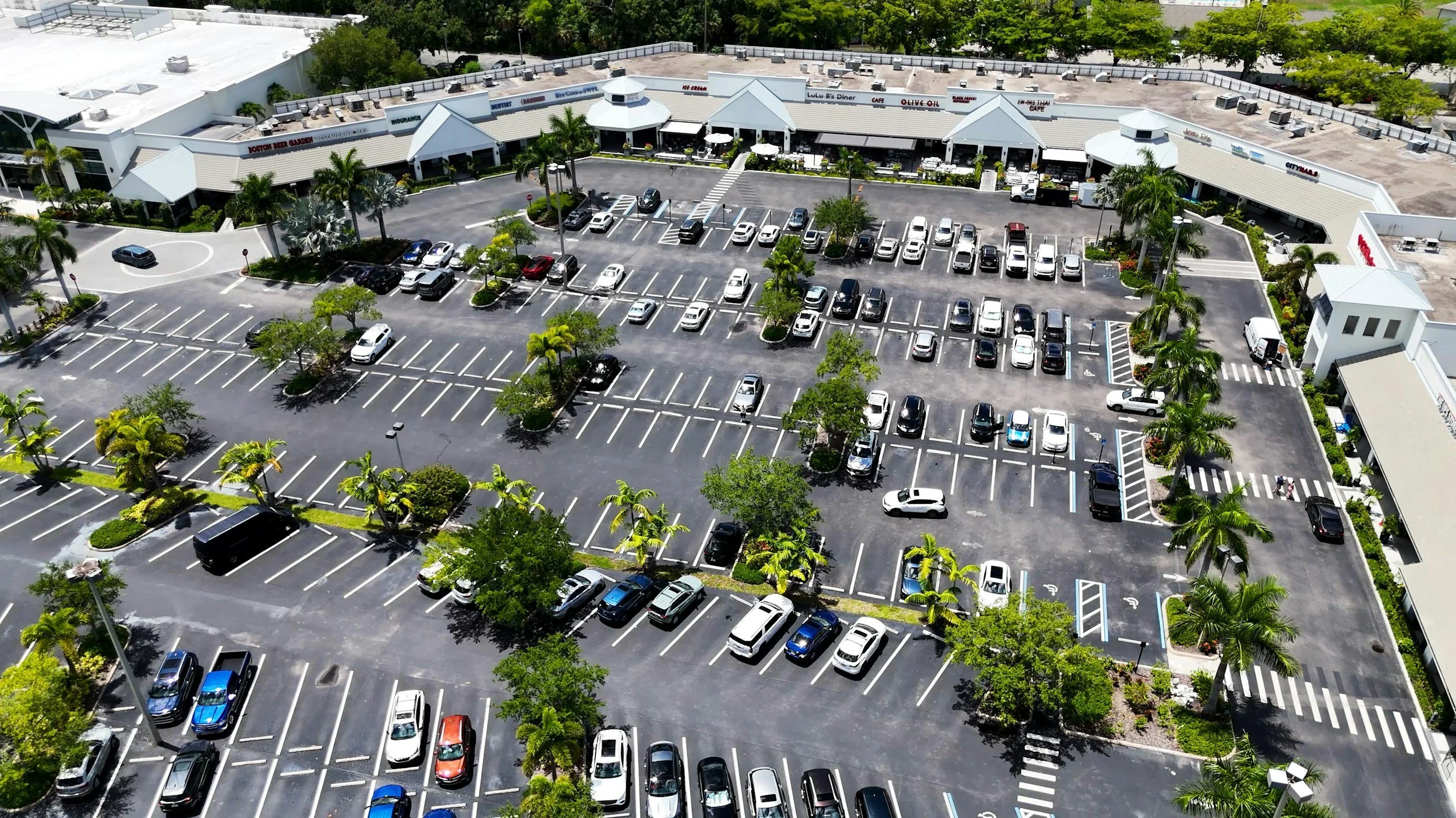 An aerial view of a large parking lot filled with white, black, and other colored cars, surrounded by trees and a shopping center with various stores and restaurants.